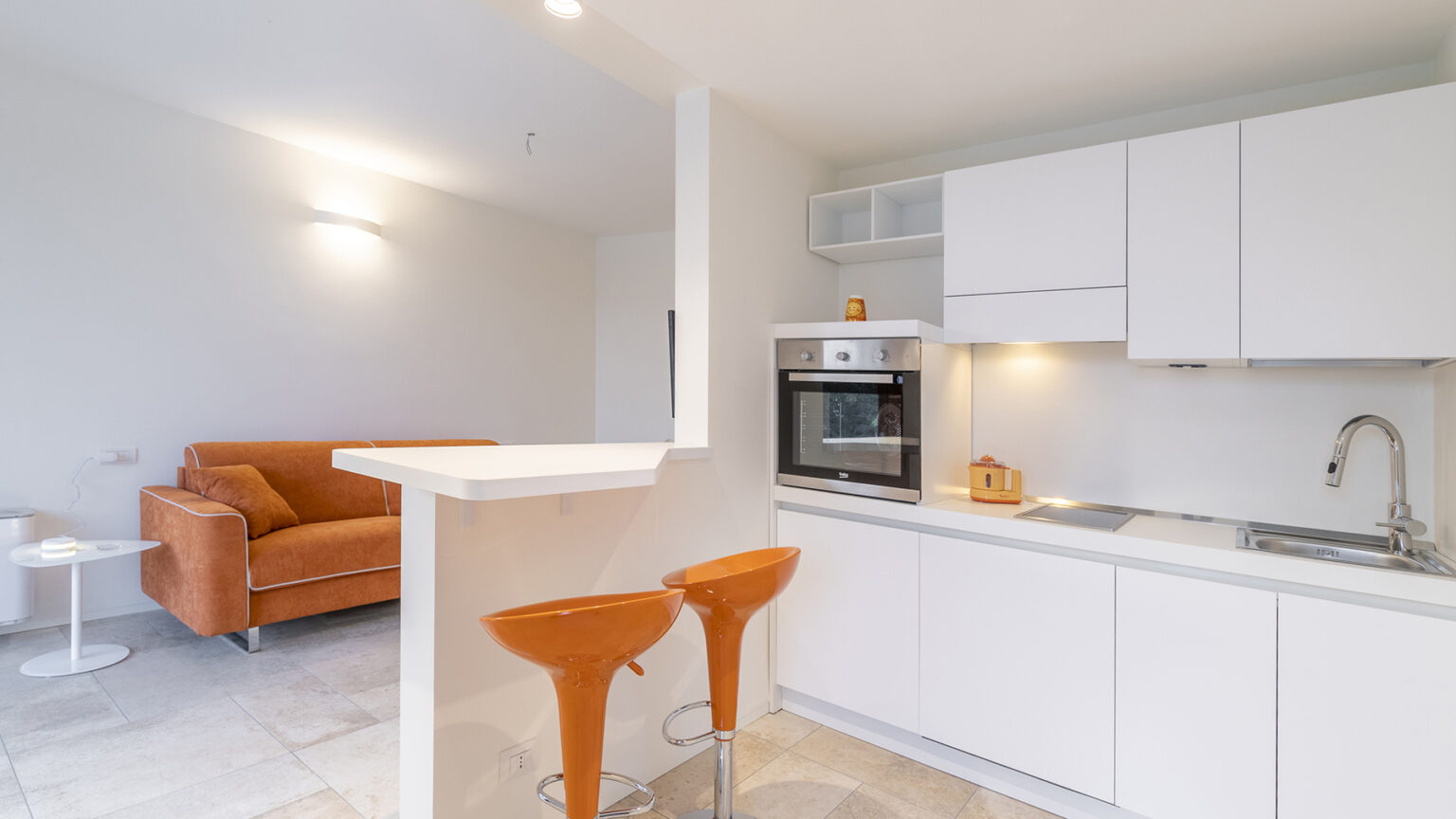 Modern kitchen and living area featuring a sleek design, white cabinetry, an oven, and two orange bar stools at a breakfast counter, with a cozy brown sofa in the background. | Only on SopranoVillas