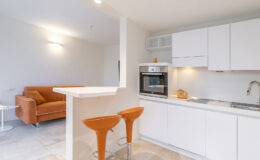 Modern kitchen and living area featuring a sleek design, white cabinetry, an oven, and two orange bar stools at a breakfast counter, with a cozy brown sofa in the background.