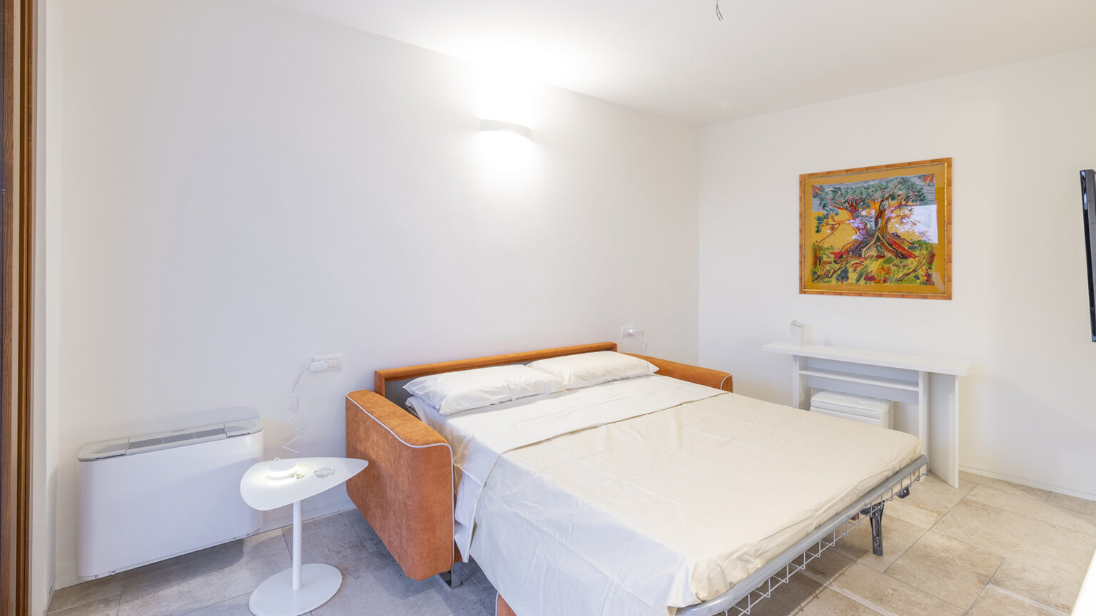 Cozy bedroom featuring a modern foldable bed with a light-colored bedspread, minimalist decor, and a colorful tree painting on the wall. The room includes a small white side table and a compact air conditioning unit, offering a bright and inviting atmosphere. | Only on SopranoVillas