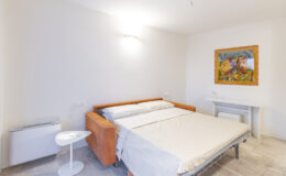 Cozy bedroom featuring a modern foldable bed with a light-colored bedspread, minimalist decor, and a colorful tree painting on the wall. The room includes a small white side table and a compact air conditioning unit, offering a bright and inviting atmosphere.