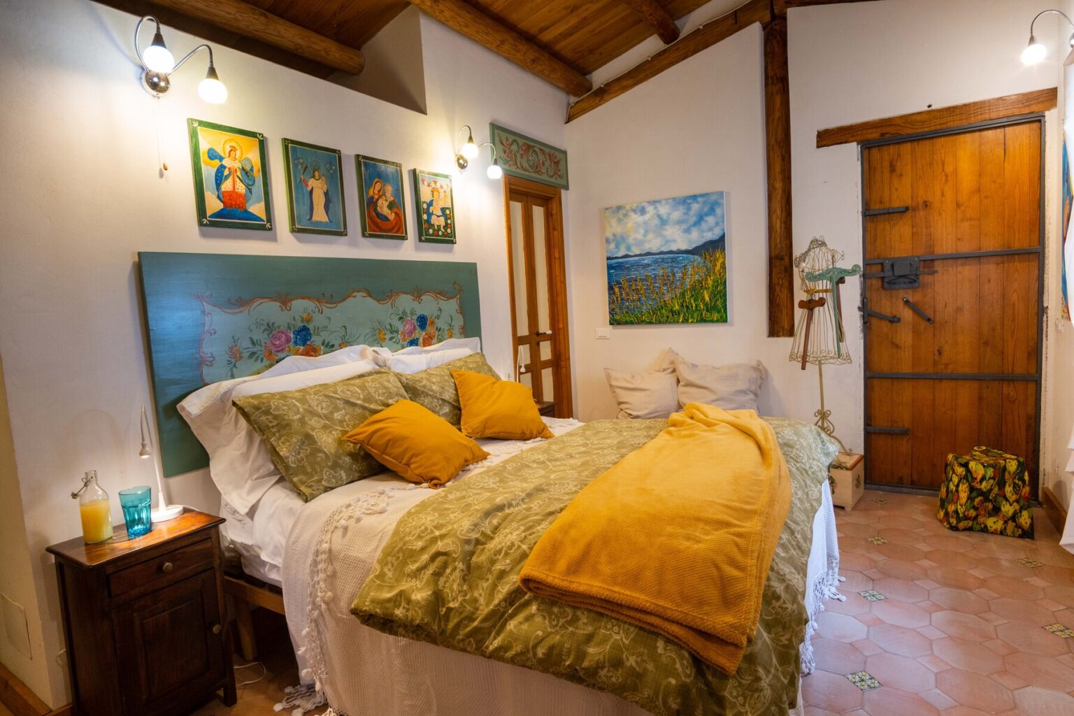 Cozy bedroom featuring a double bed with decorative pillows, a colorful headboard, and artwork on the walls. The room has wooden accents, warm lighting, and a rustic wooden door, creating a welcoming atmosphere. | Only on SopranoVillas