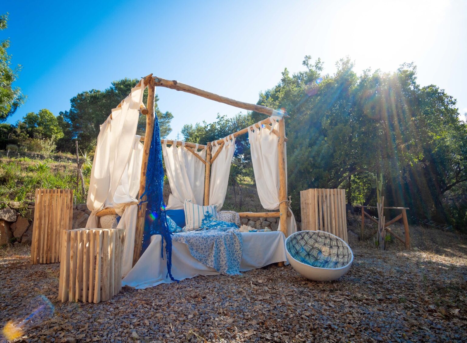 Outdoor lounge area with a wooden pergola draped in white fabric and blue accents, surrounded by greenery and sunlight, featuring a cozy seating arrangement and decorative elements. | Only on SopranoVillas