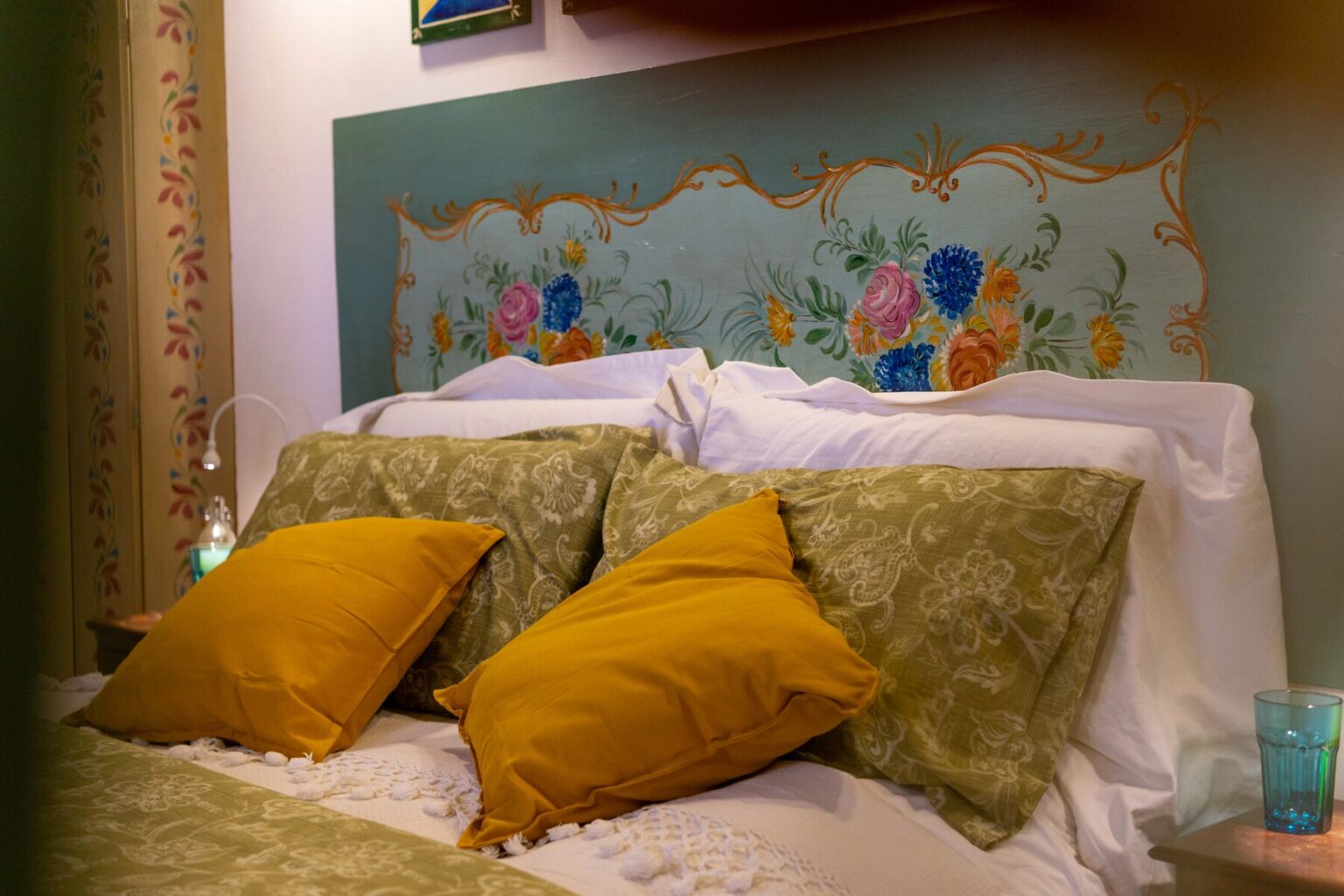 Cozy bedroom featuring a beautifully painted headboard with floral designs, complemented by green and mustard yellow decorative pillows on a neatly made bed. Soft lighting and a decorative lamp add warmth to the inviting atmosphere. | Only on SopranoVillas