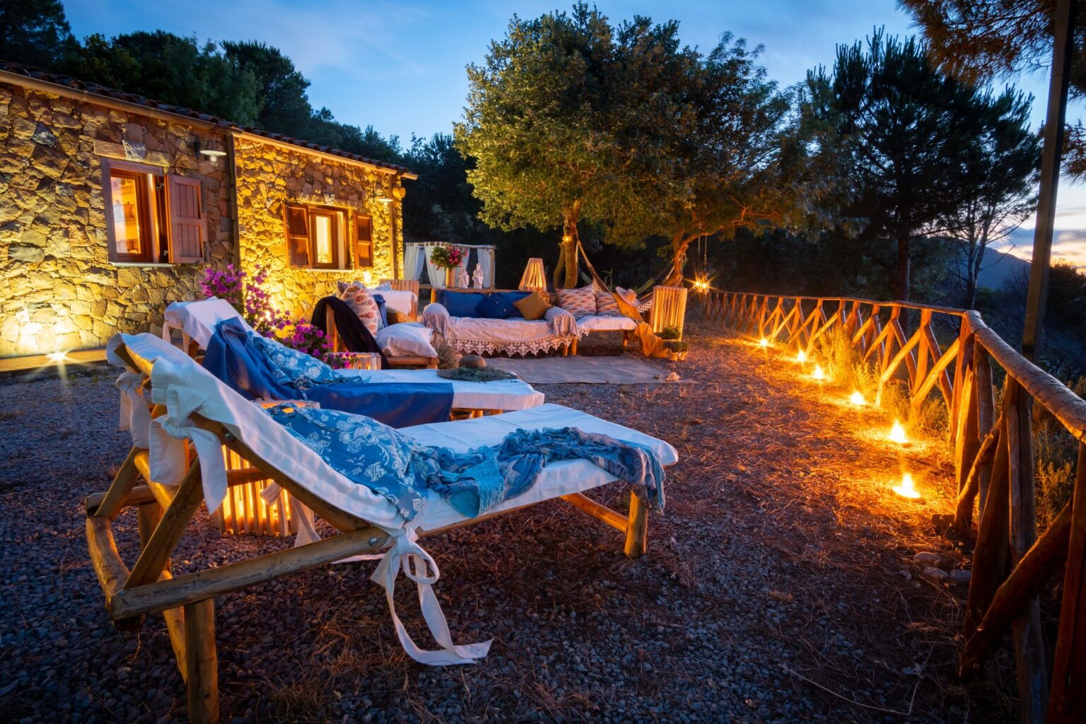 Cozy outdoor seating area with lounge chairs and decorative blankets, illuminated by soft lighting at dusk, set against a rustic stone house surrounded by trees. | Only on SopranoVillas