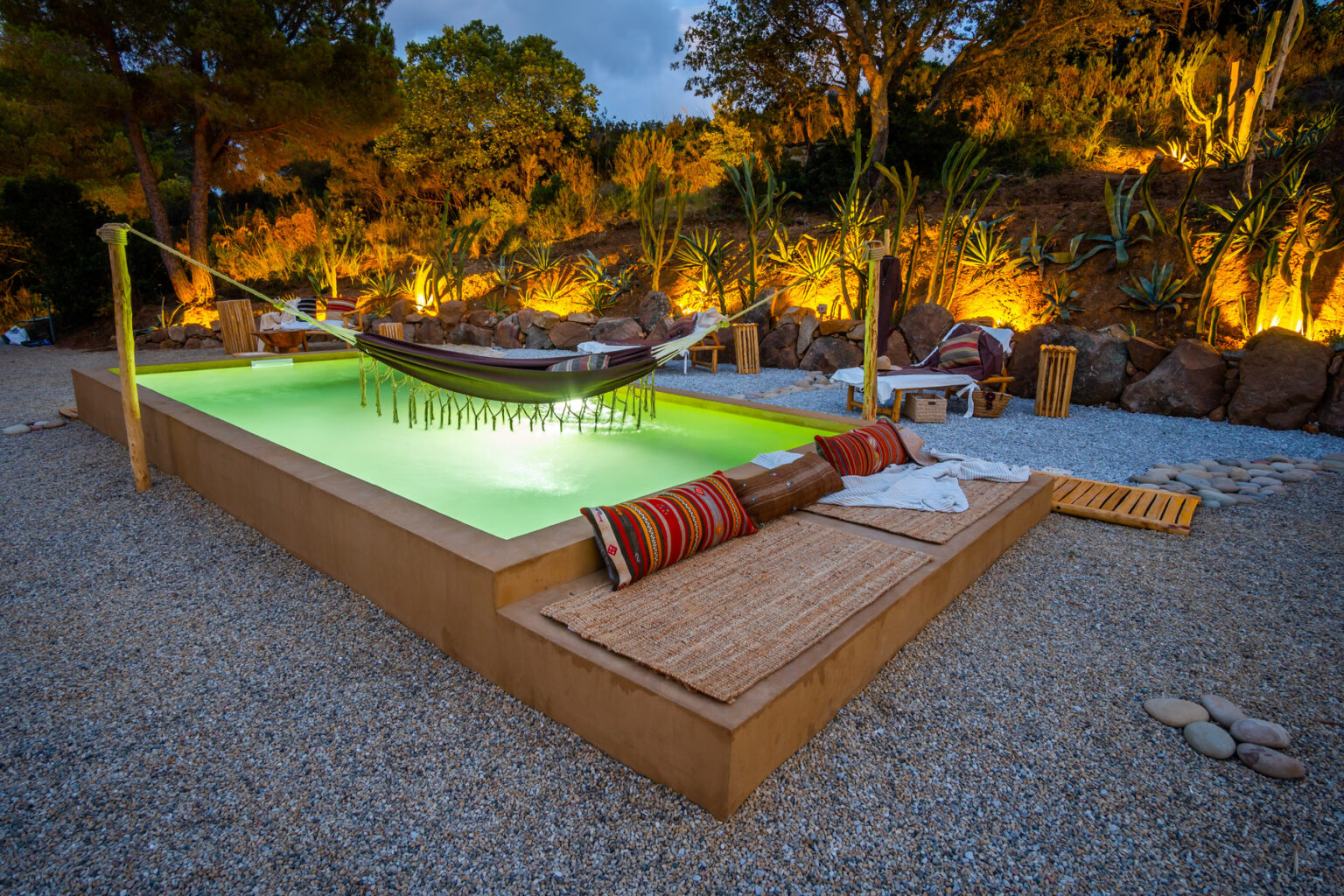 Serene outdoor space featuring a modern green-lit pool surrounded by gravel, a cozy hammock, decorative pillows, and ambient lighting from surrounding plants, creating a relaxing atmosphere for leisure and tranquility. | Only on SopranoVillas