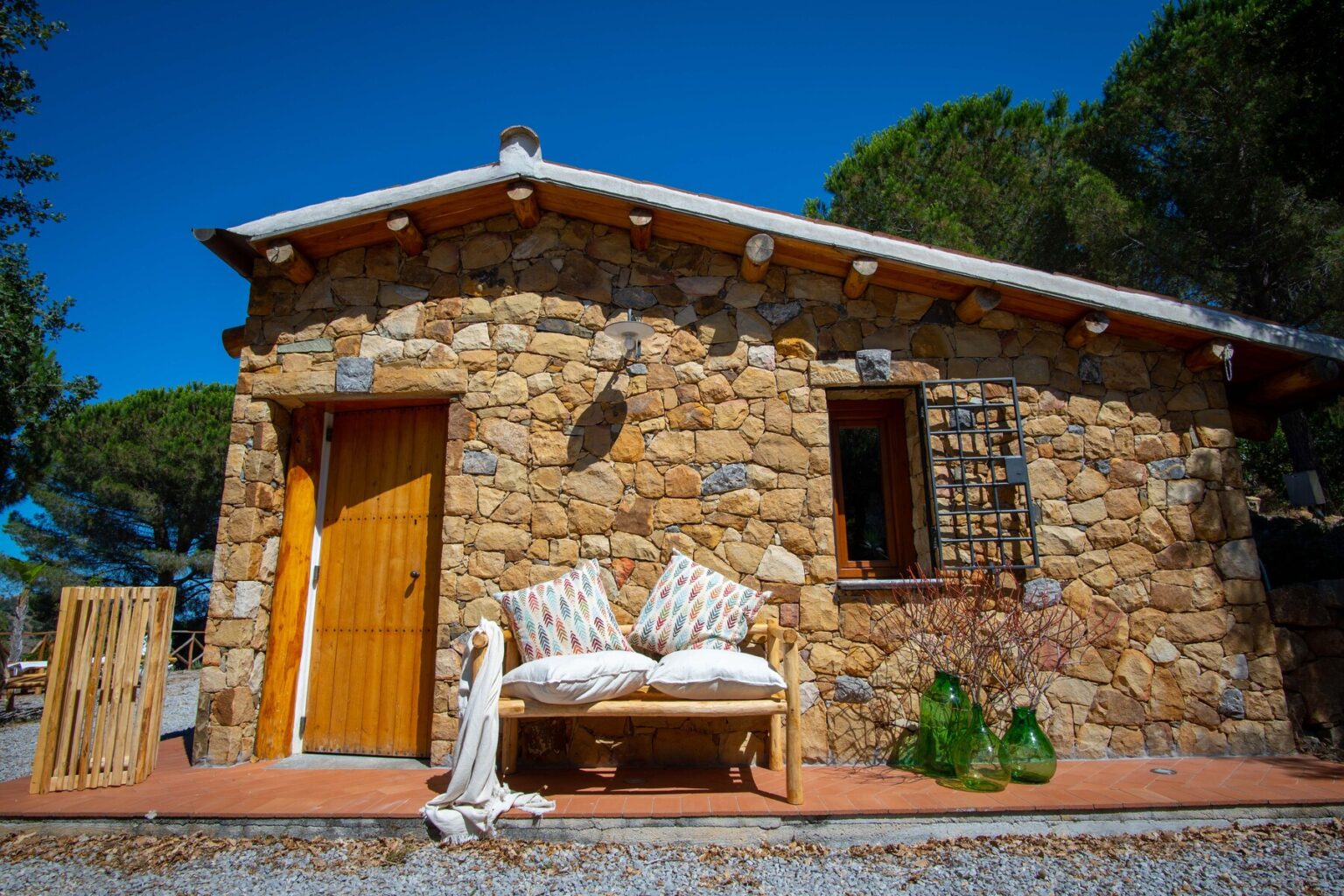 Charming stone cottage with a wooden door and a cozy outdoor seating area, surrounded by greenery and blue skies, perfect for a tranquil getaway. | Only on SopranoVillas