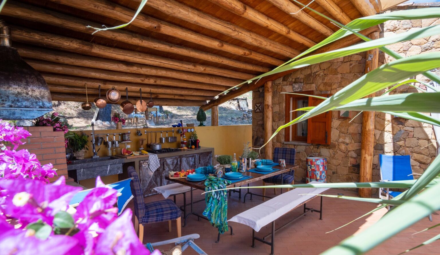 Cozy outdoor dining area with wooden beams, stone walls, and colorful decor, featuring a table set for a meal, surrounded by lush greenery and vibrant flowers. Ideal for entertaining in a rustic setting. | Only on SopranoVillas