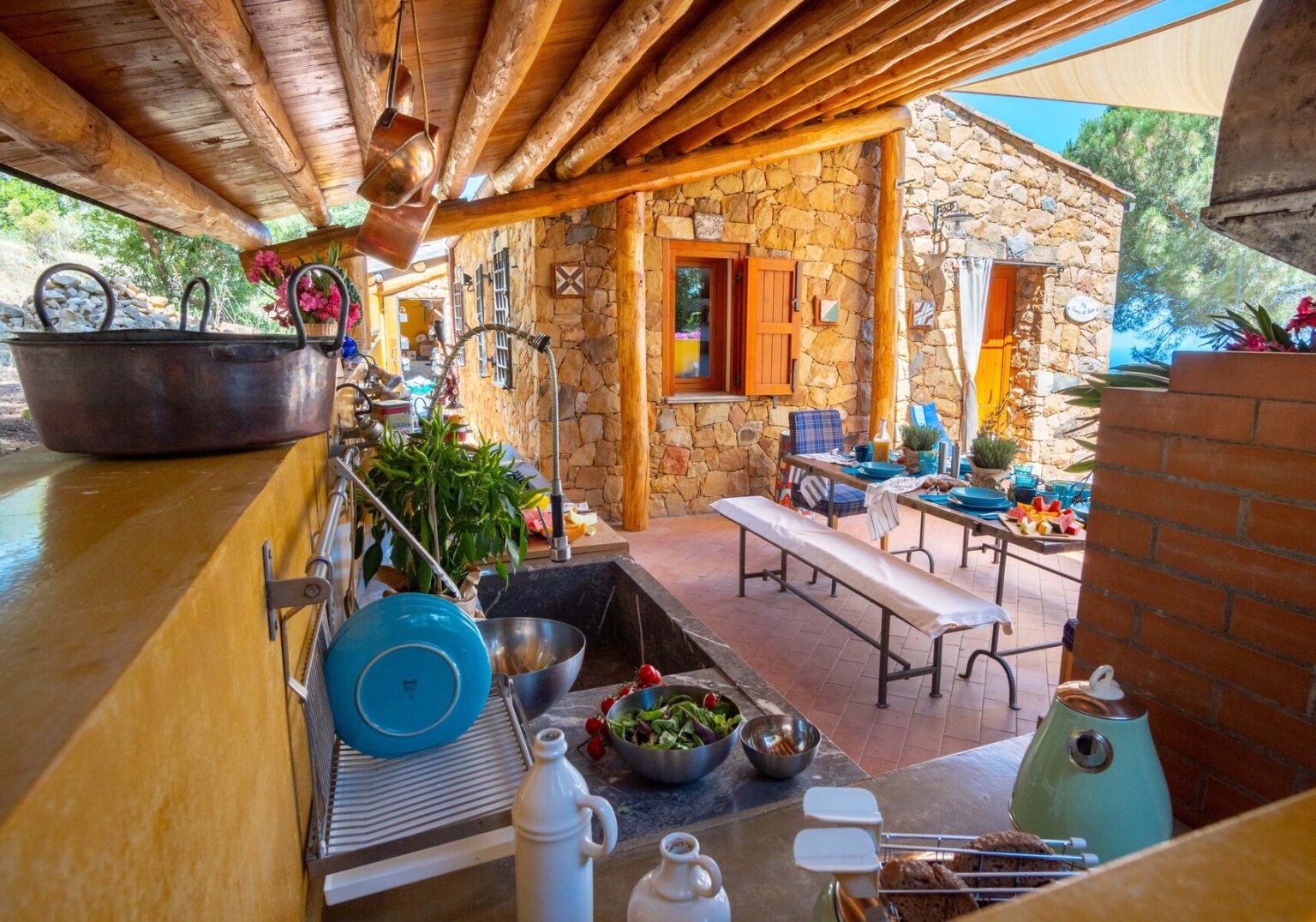 Cozy outdoor kitchen with stone walls, wooden beams, and a rustic dining area featuring a long table, surrounded by greenery and kitchenware, ideal for al fresco dining and gatherings. | Only on SopranoVillas