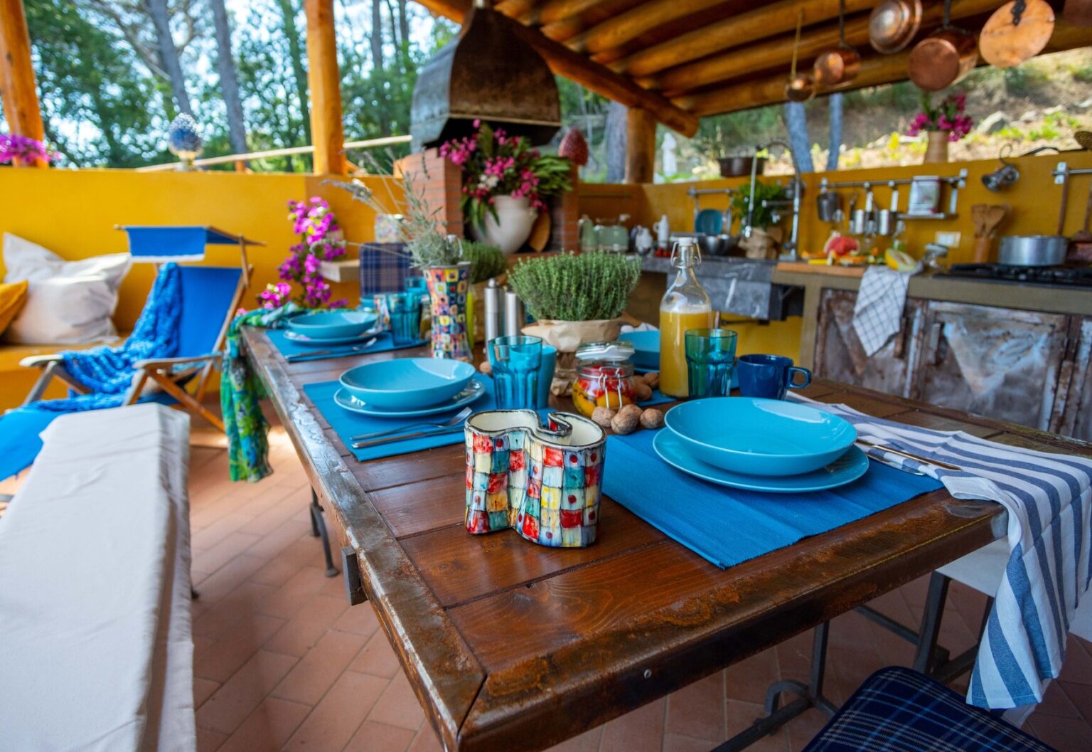 Vibrant outdoor dining setup featuring a wooden table with blue plates, colorful mugs, and fresh greenery, surrounded by a bright yellow wall and decorative plants, ideal for a summer gathering. | Only on SopranoVillas
