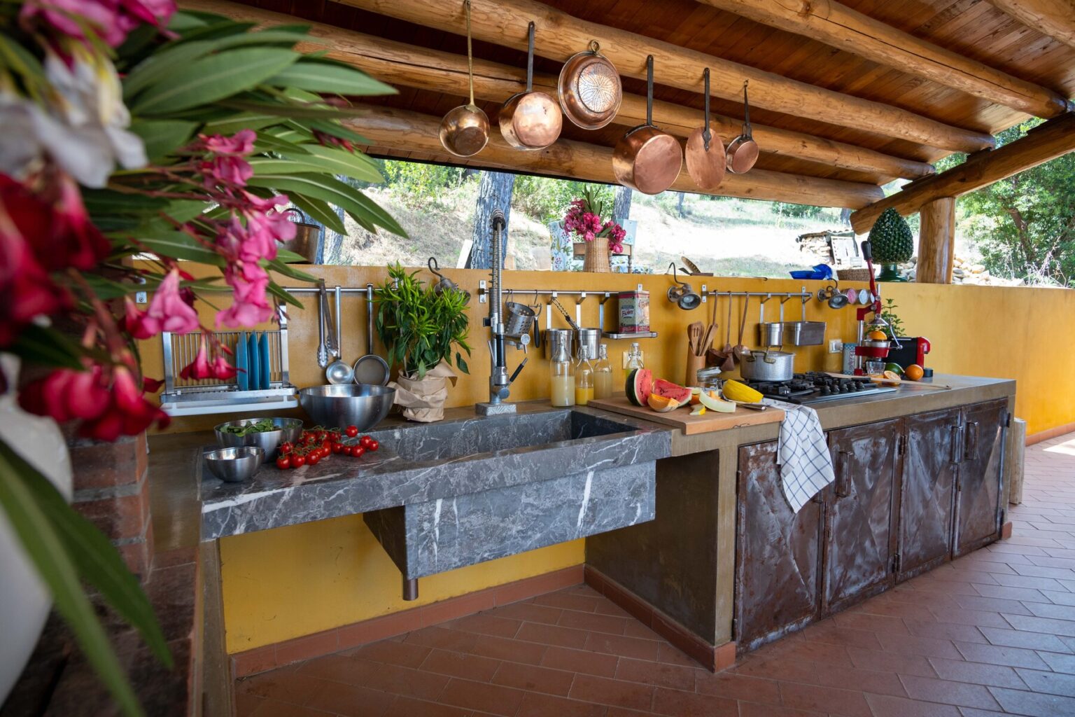 Outdoor kitchen with a rustic design featuring a stone sink, hanging copper pots, vibrant flowers, and a variety of cooking utensils, set against a yellow wall and wooden beams. | Only on SopranoVillas