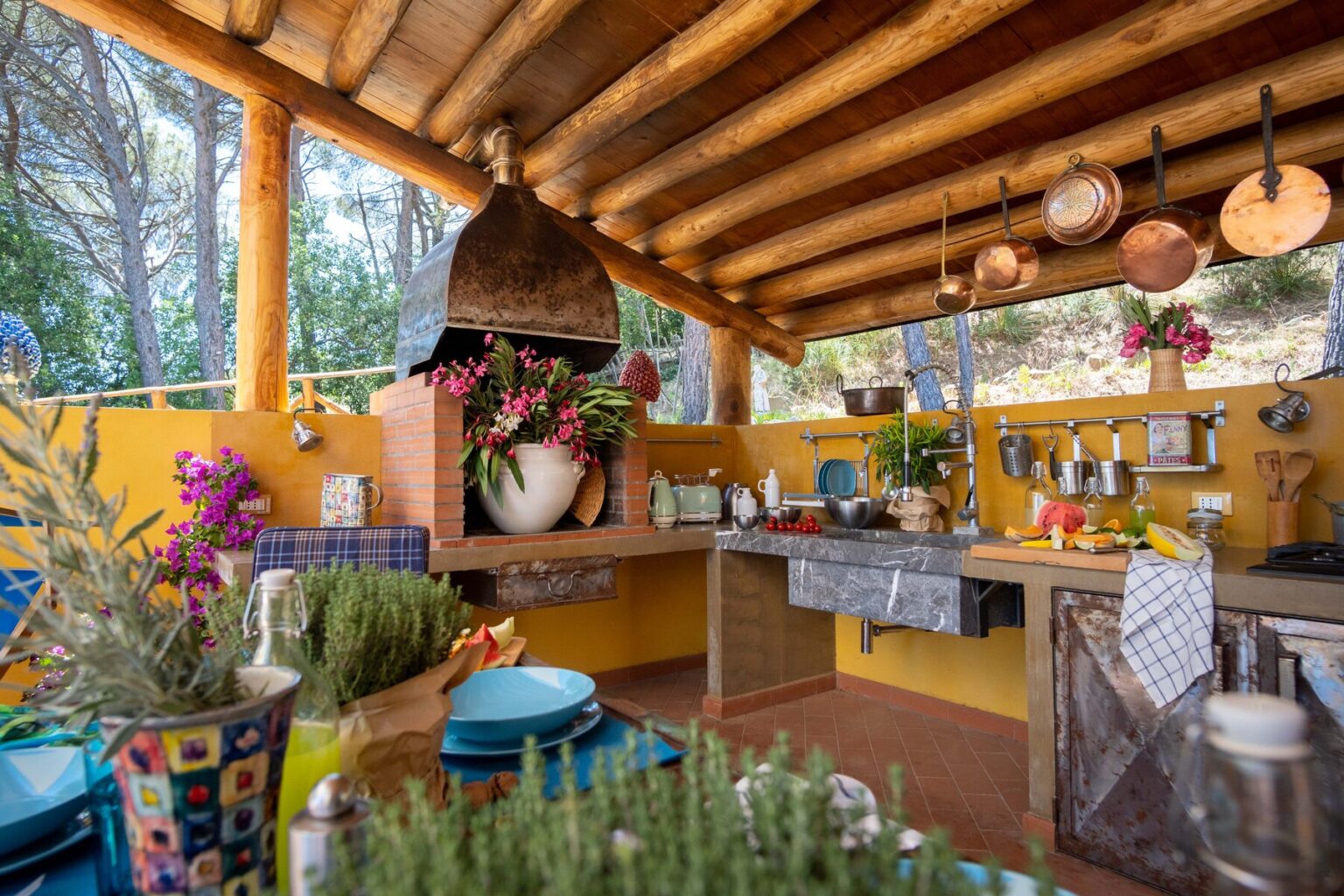 Cozy outdoor kitchen featuring rustic wooden beams, colorful pottery, and vibrant flowers. The space is adorned with hanging copper pots and a stone countertop, creating an inviting atmosphere for cooking and entertaining. | Only on SopranoVillas