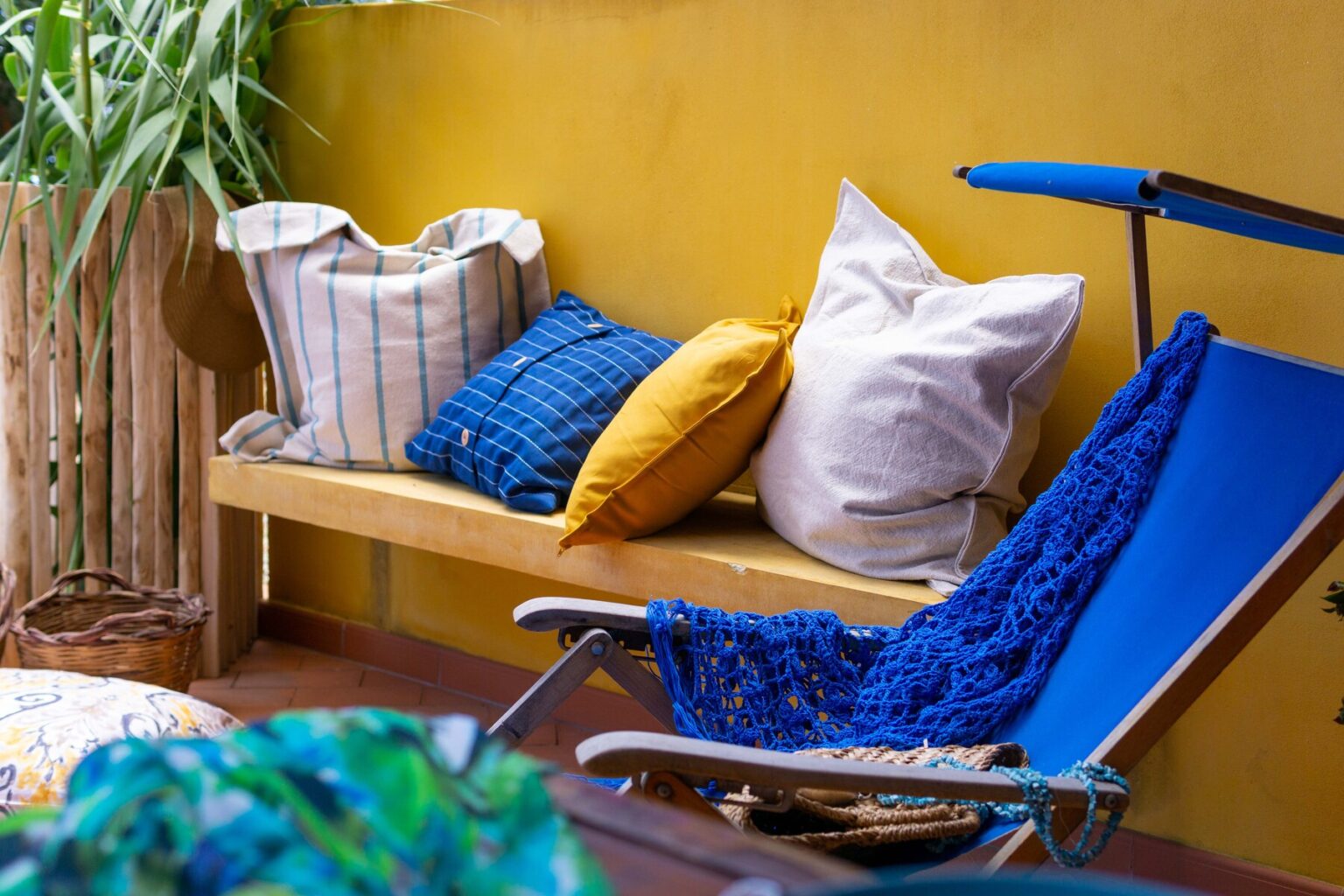 Cozy outdoor seating area featuring colorful cushions on a wooden bench and a blue lounge chair, surrounded by greenery against a vibrant yellow wall. | Only on SopranoVillas
