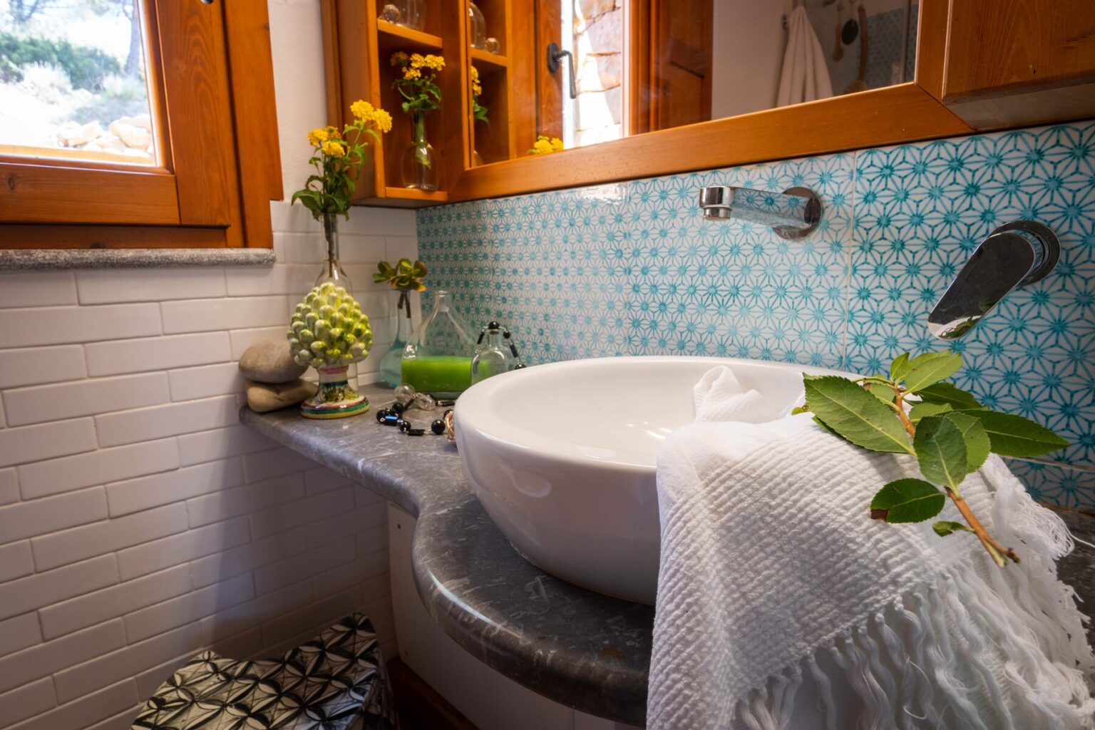 Modern bathroom design featuring a round white sink, natural elements like plants and flowers, and vibrant blue patterned tiles. The space is illuminated by natural light from a window, enhancing the serene atmosphere. | Only on SopranoVillas