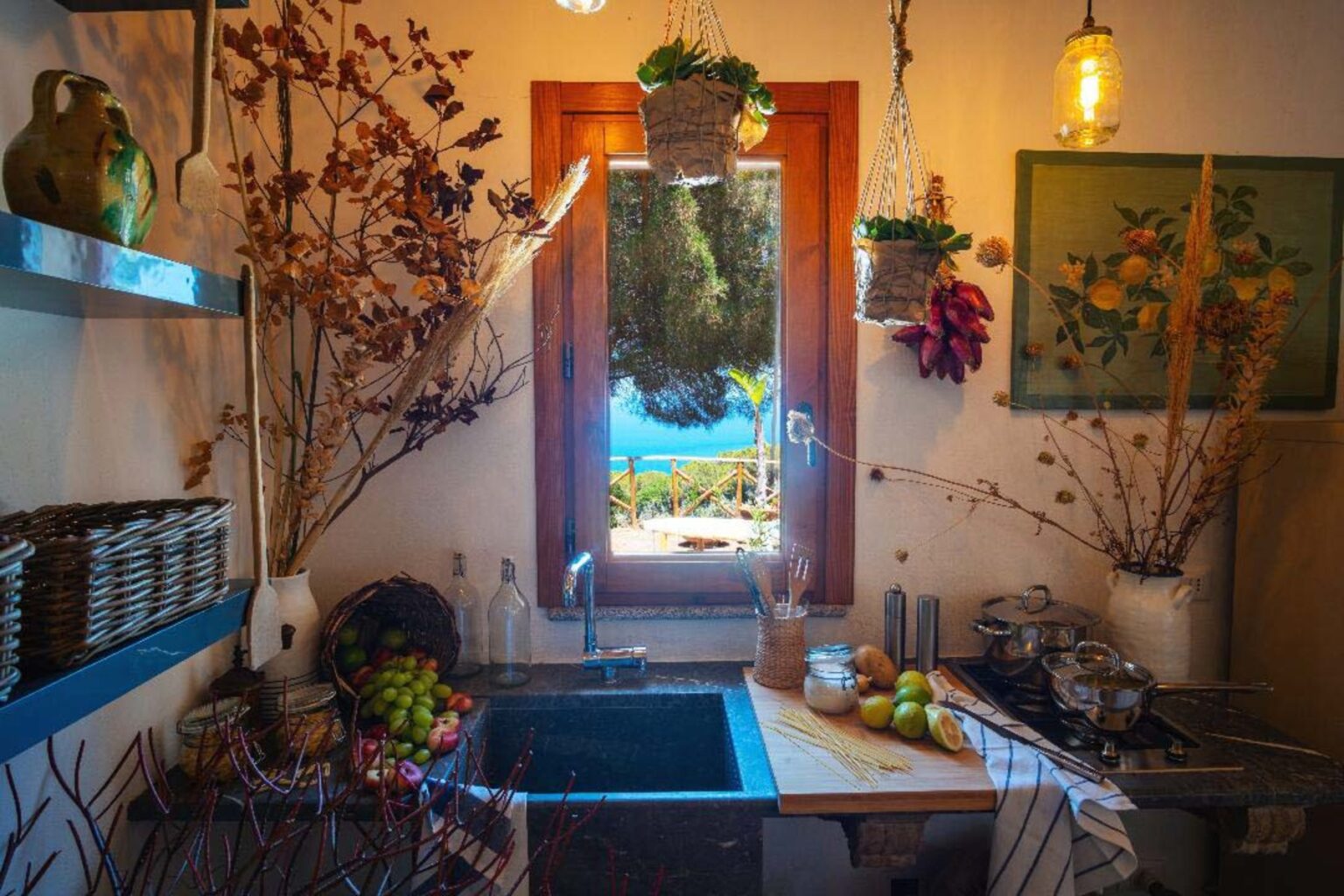 Cozy kitchen with blue shelves, a sink, and decorative plants. Sunlight streams through a window showcasing a vibrant outdoor view. Rustic decor includes dried flowers and fruit, creating a warm and inviting atmosphere. | Only on SopranoVillas