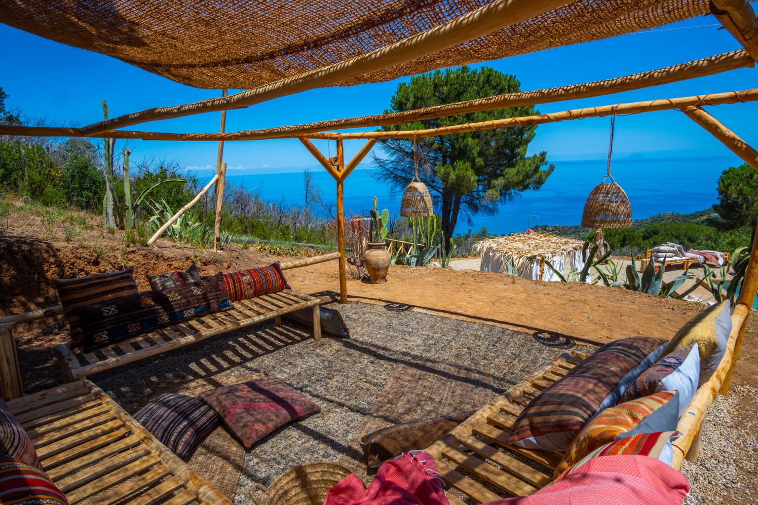 Cozy outdoor seating area with bamboo furniture and colorful cushions, overlooking a scenic view of the ocean and greenery under a woven shade structure. Ideal for relaxation and enjoying nature. | Only on SopranoVillas