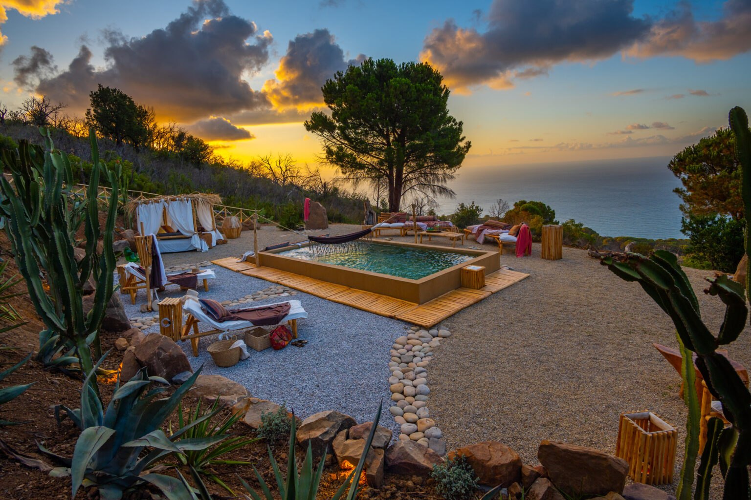 Scenic outdoor pool area at sunset, surrounded by lush greenery and desert plants, offering a tranquil view of the ocean. Cozy seating arrangements enhance the relaxing atmosphere, ideal for leisure and meditation. | Only on SopranoVillas