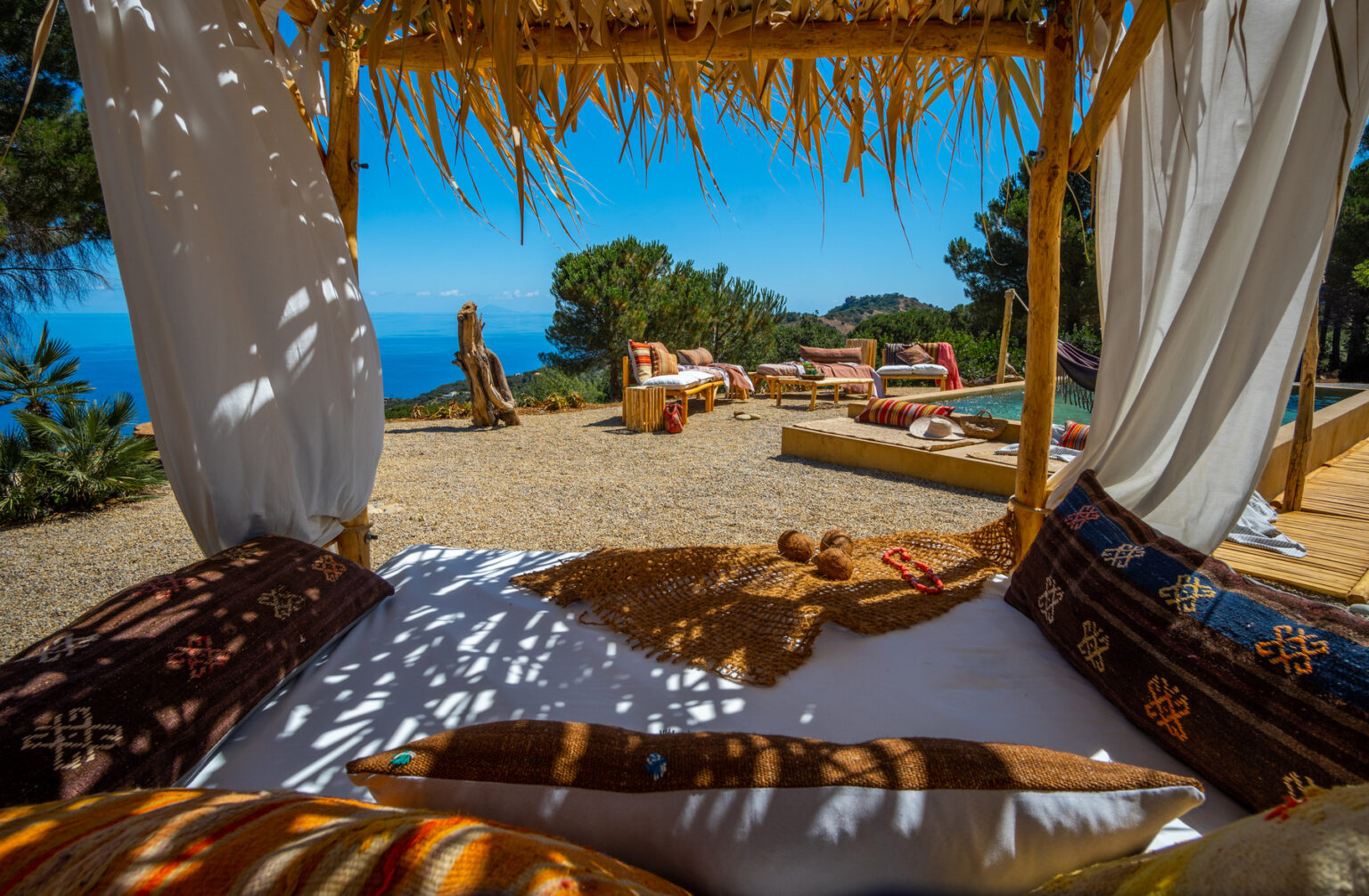 Relaxing outdoor lounge area with a view of the ocean, featuring a thatched gazebo, colorful cushions, and comfortable seating. The setting includes a wooden deck, a pool, and lush greenery, creating a serene atmosphere for leisure and tranquility. | Only on SopranoVillas