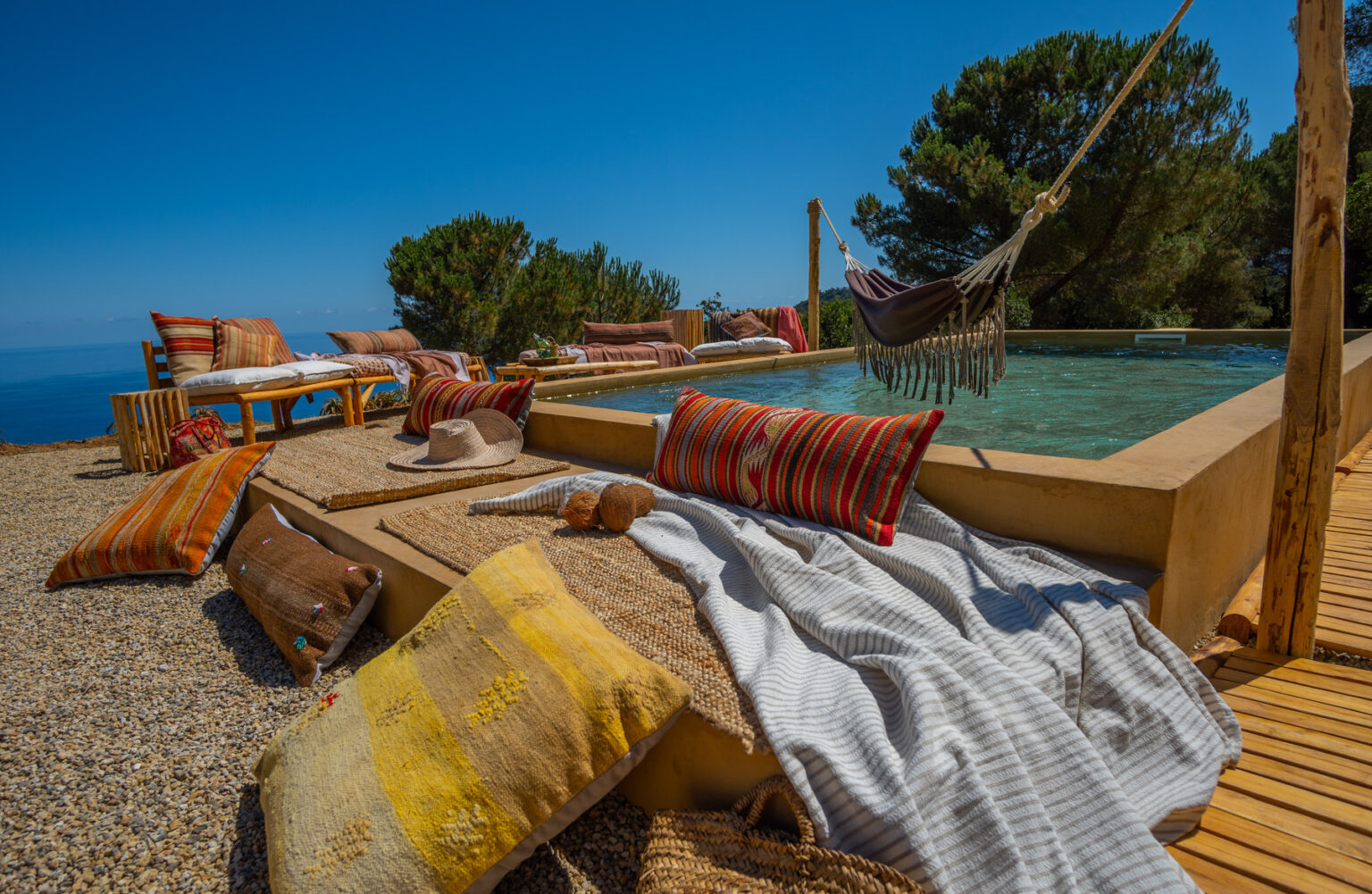 Relaxing outdoor space featuring a pool, colorful cushions, and a hammock with a scenic ocean view. The area is adorned with textured rugs, sun hats, and decorative throws, creating a cozy and inviting atmosphere. | Only on SopranoVillas