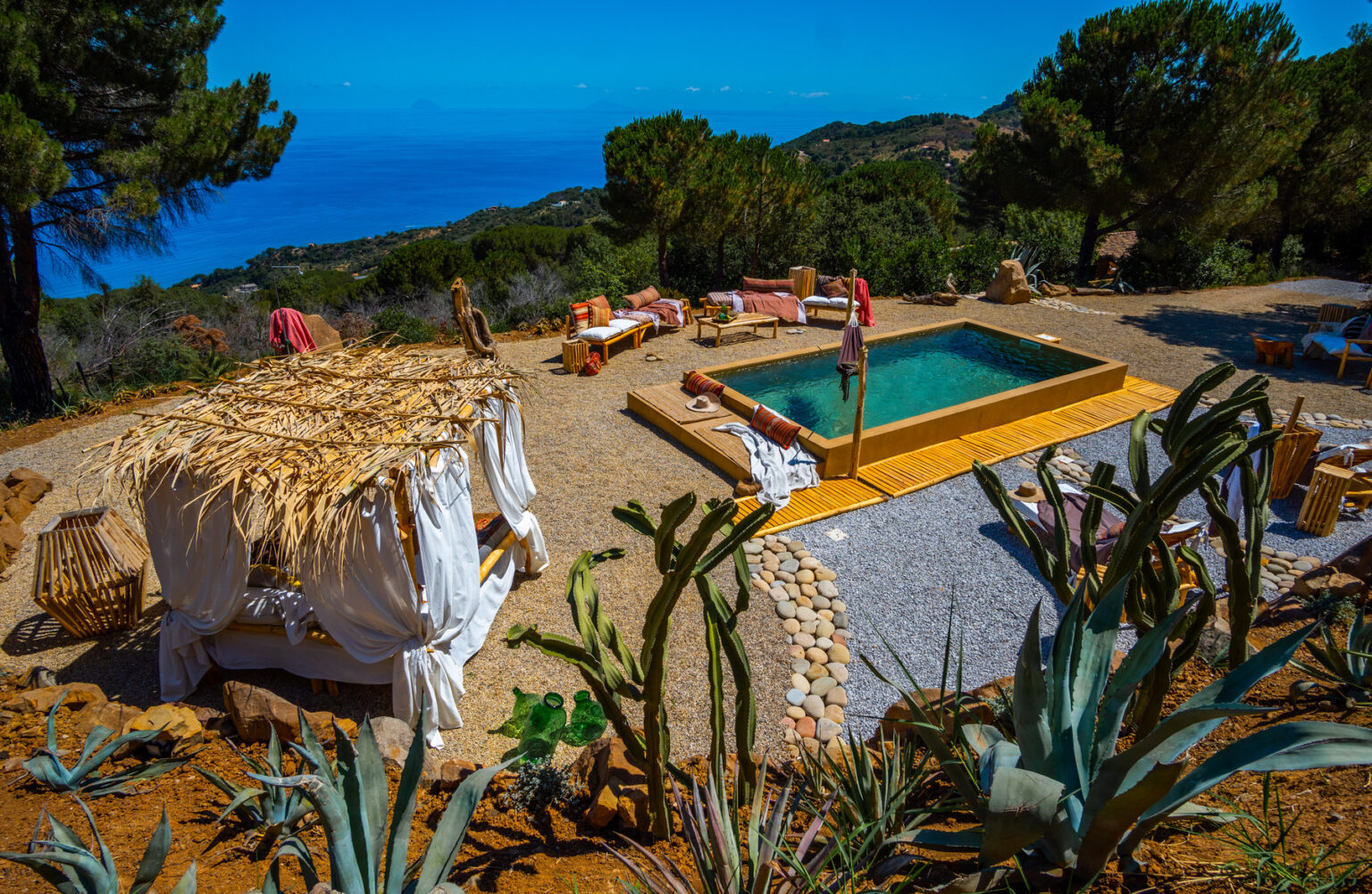 Scenic outdoor pool area with straw cabanas, surrounded by lush greenery and cacti, overlooking a tranquil blue ocean and hills. Ideal for relaxation and leisure. | Only on SopranoVillas