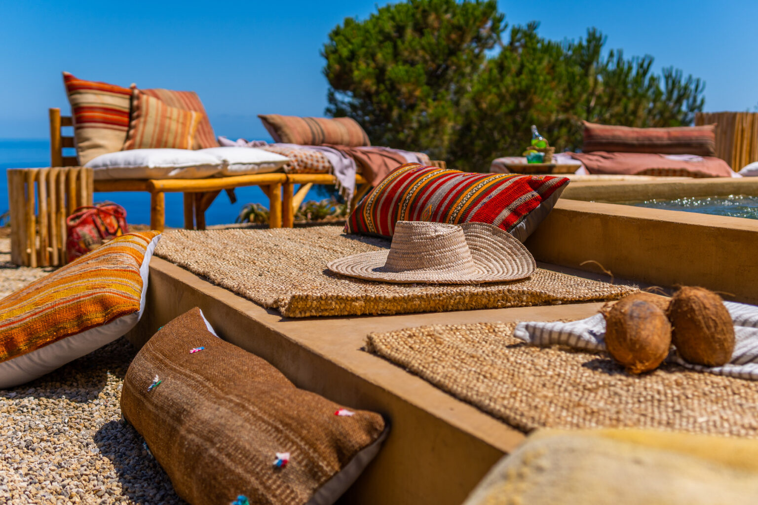 Cozy outdoor lounge area featuring woven rugs, decorative pillows, and a straw hat, set against a serene ocean backdrop and lush greenery. Perfect for relaxation and summer gatherings. | Only on SopranoVillas