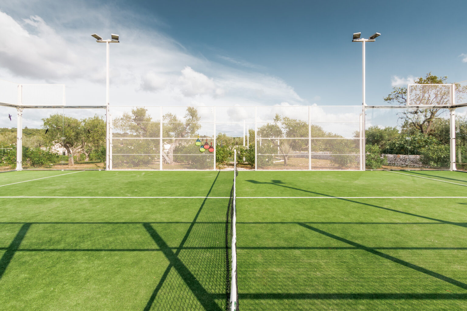 A panoramic view of a well-maintained artificial turf sports field with goalposts, surrounded by greenery and under a clear blue sky, ideal for soccer or other field sports. | Only on SopranoVillas