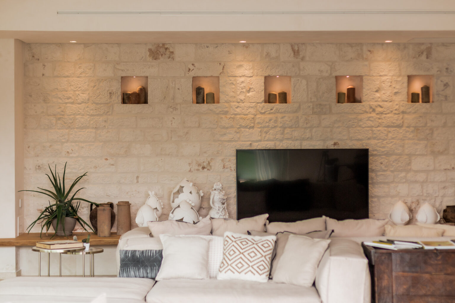 Modern living room featuring a textured stone wall with illuminated niches displaying candles, a large flat-screen TV, and a comfortable sofa adorned with decorative pillows. A stylish plant and decorative items complete the cozy atmosphere. | Only on SopranoVillas