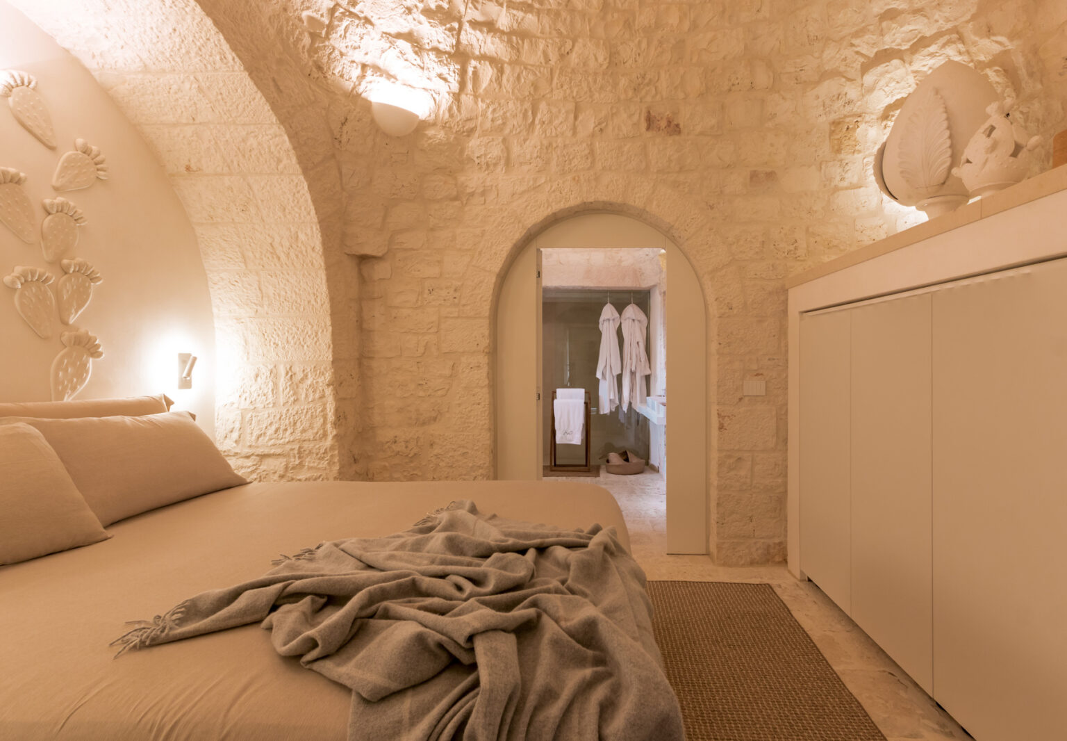 A cozy bedroom featuring arched stone walls, a minimalist design with a large bed covered in a soft blanket, and a glimpse of a stylish bathroom through an open doorway. Warm lighting enhances the serene atmosphere, perfect for relaxation. | Only on SopranoVillas