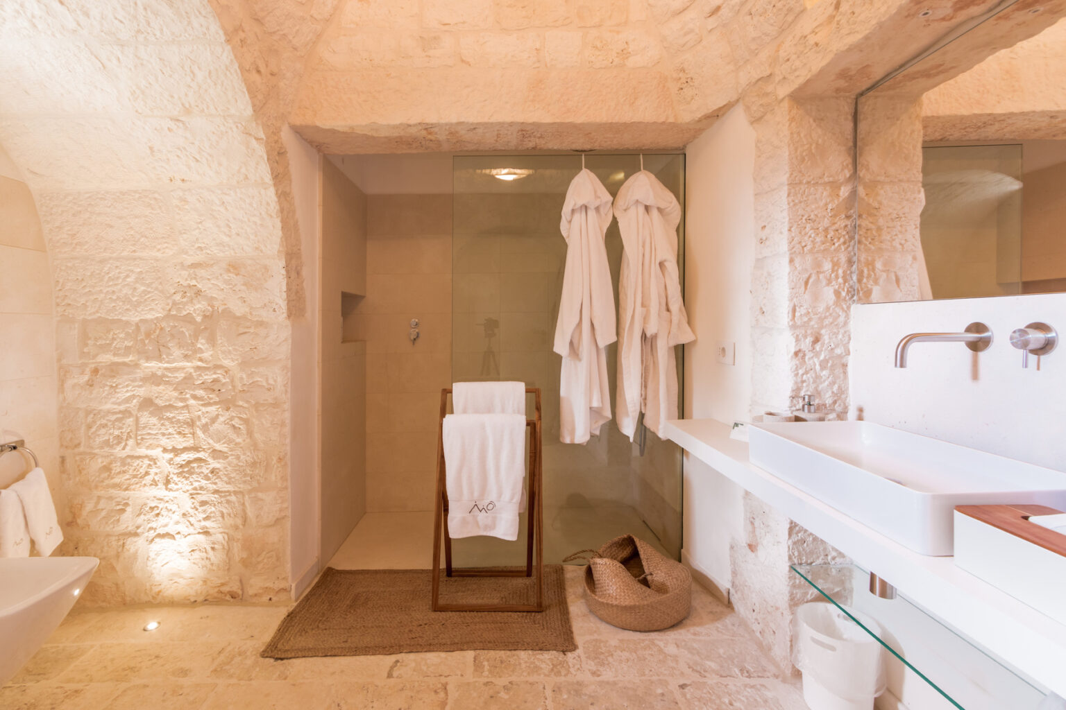 Modern bathroom interior featuring a stone wall, a glass shower, plush white bathrobes hanging, and a stylish sink area with wooden accents. Soft lighting enhances the serene atmosphere, complemented by a woven rug and decorative basket. Ideal for luxury home or hotel design inspiration. | Only on SopranoVillas