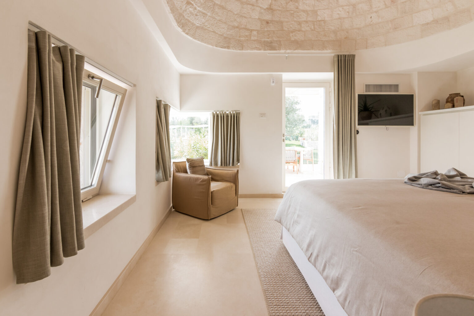 Modern bedroom featuring a cozy bed, natural light from large windows, minimalist decor, and comfortable seating area with a stylish armchair. The soft color palette and textured ceiling create a serene atmosphere. | Only on SopranoVillas