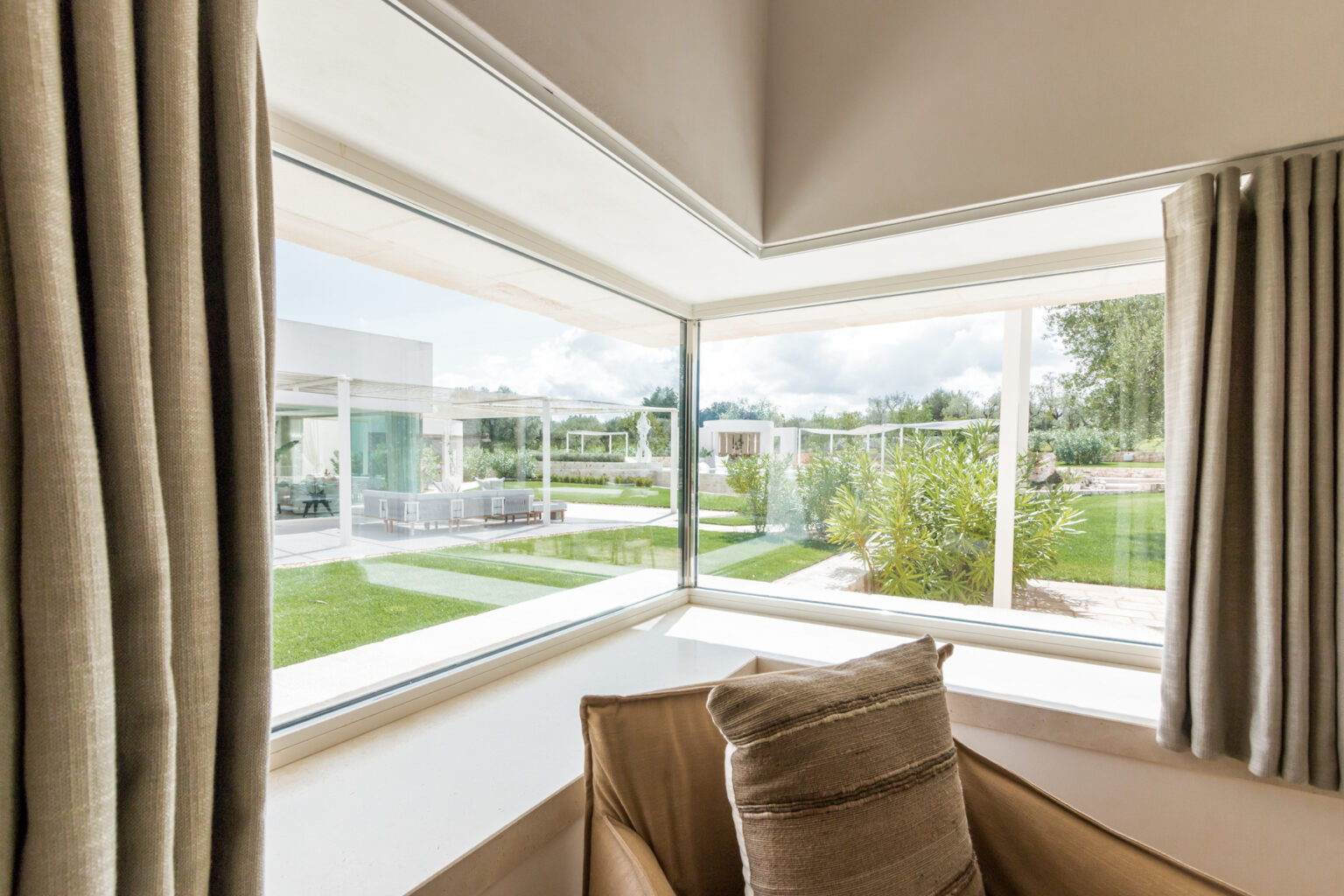 Cozy corner of a modern living room with large windows showcasing a lush garden view, featuring beige curtains and a comfortable armchair with decorative pillows. | Only on SopranoVillas
