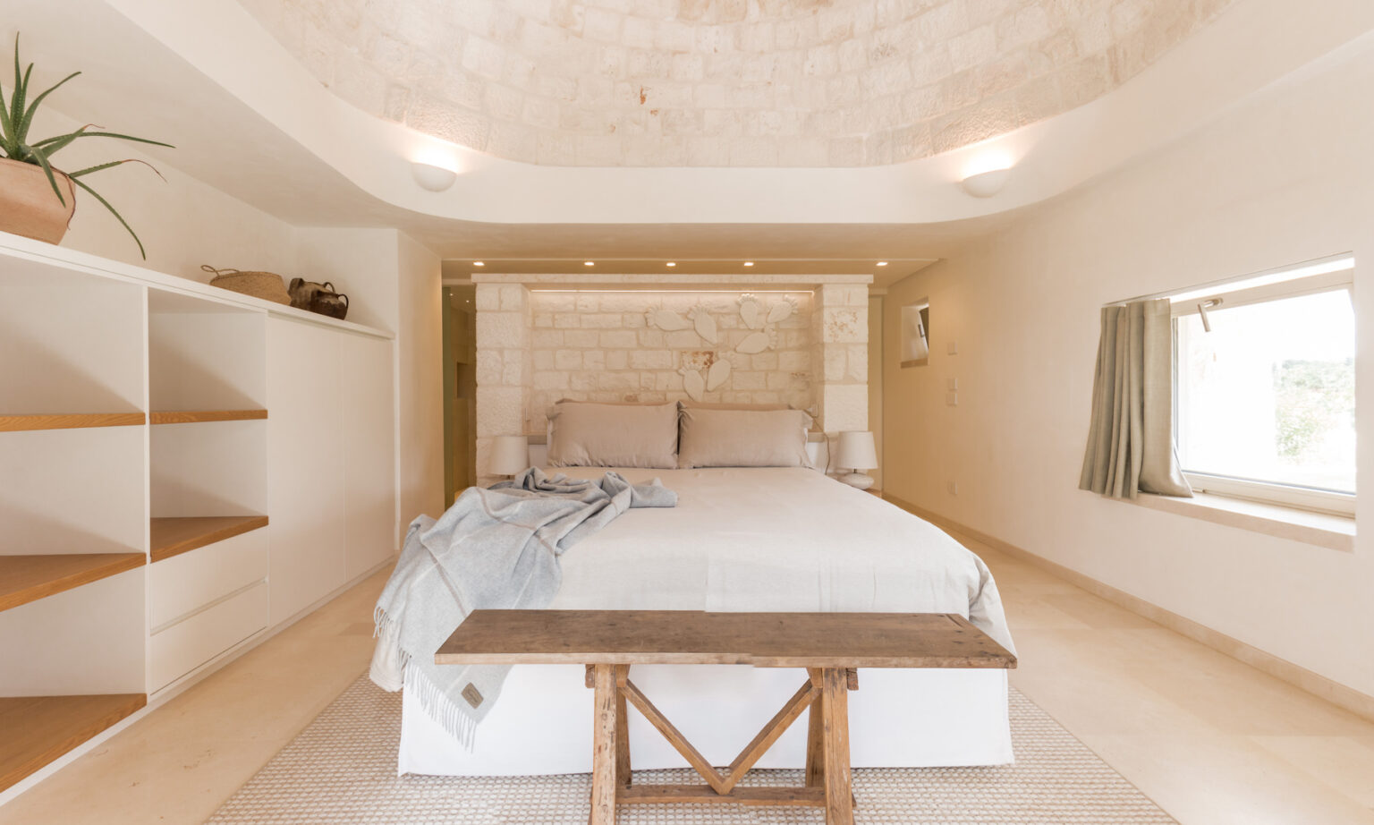Modern bedroom interior featuring a king-sized bed with neutral bedding, a wooden bench, and minimalist decor. The room has a unique stone wall backdrop, soft lighting, and a large window with curtains, creating a serene and inviting atmosphere. | Only on SopranoVillas