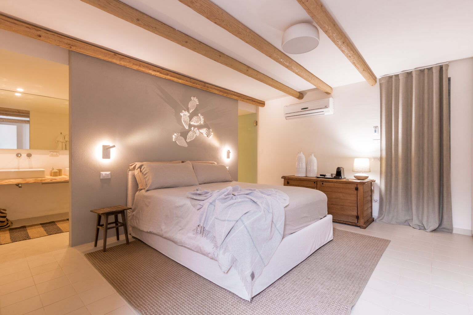 Modern bedroom interior featuring a cozy bed with light-colored bedding, wooden accents, and decorative wall art. The room includes soft lighting, a bedside table with a lamp, and a stylish curtain, creating a warm and inviting atmosphere. | Only on SopranoVillas