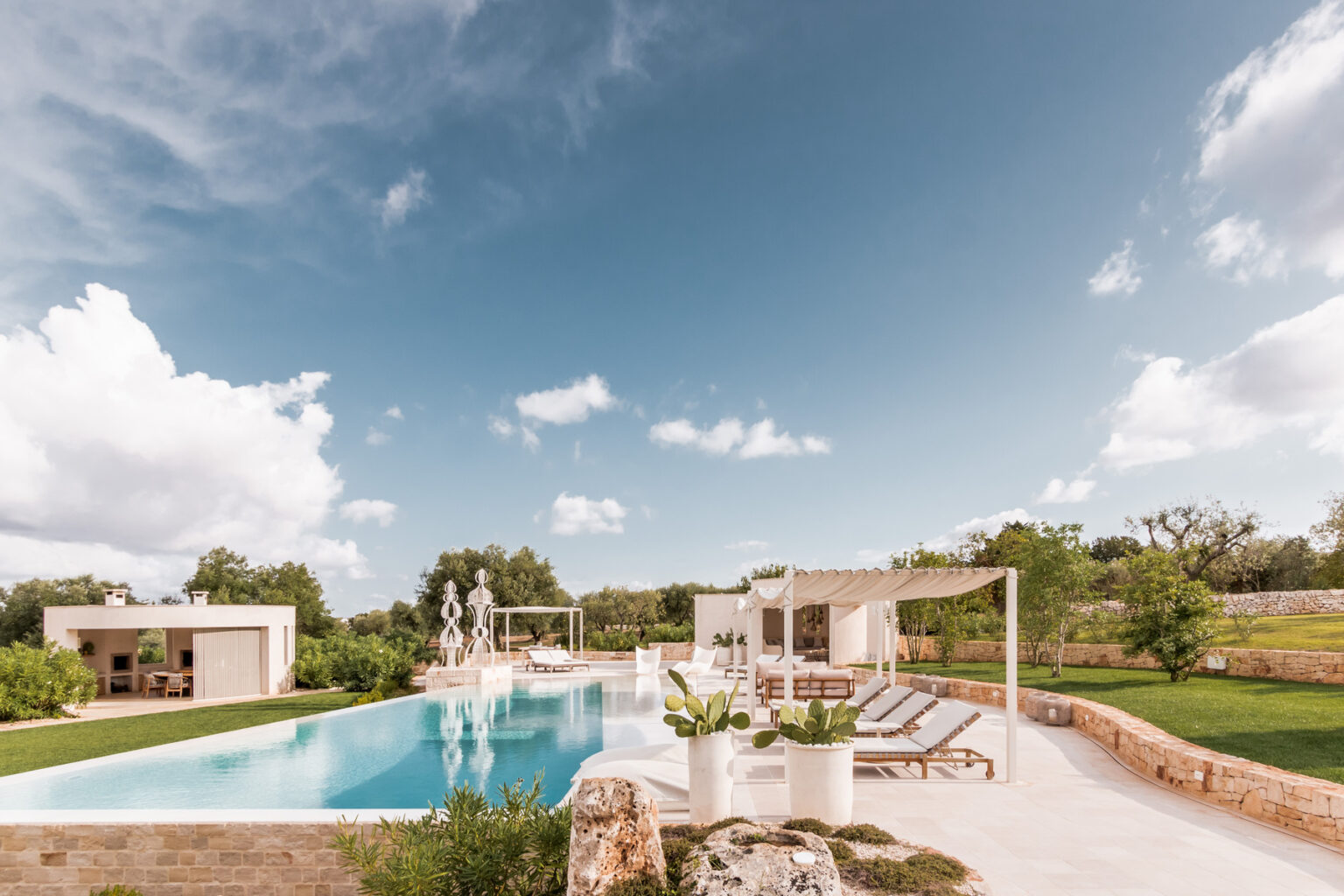 Modern villa with a spacious outdoor pool surrounded by lush greenery, sun loungers, and a pergola, under a clear blue sky. Ideal for luxury relaxation and outdoor gatherings. | Only on SopranoVillas