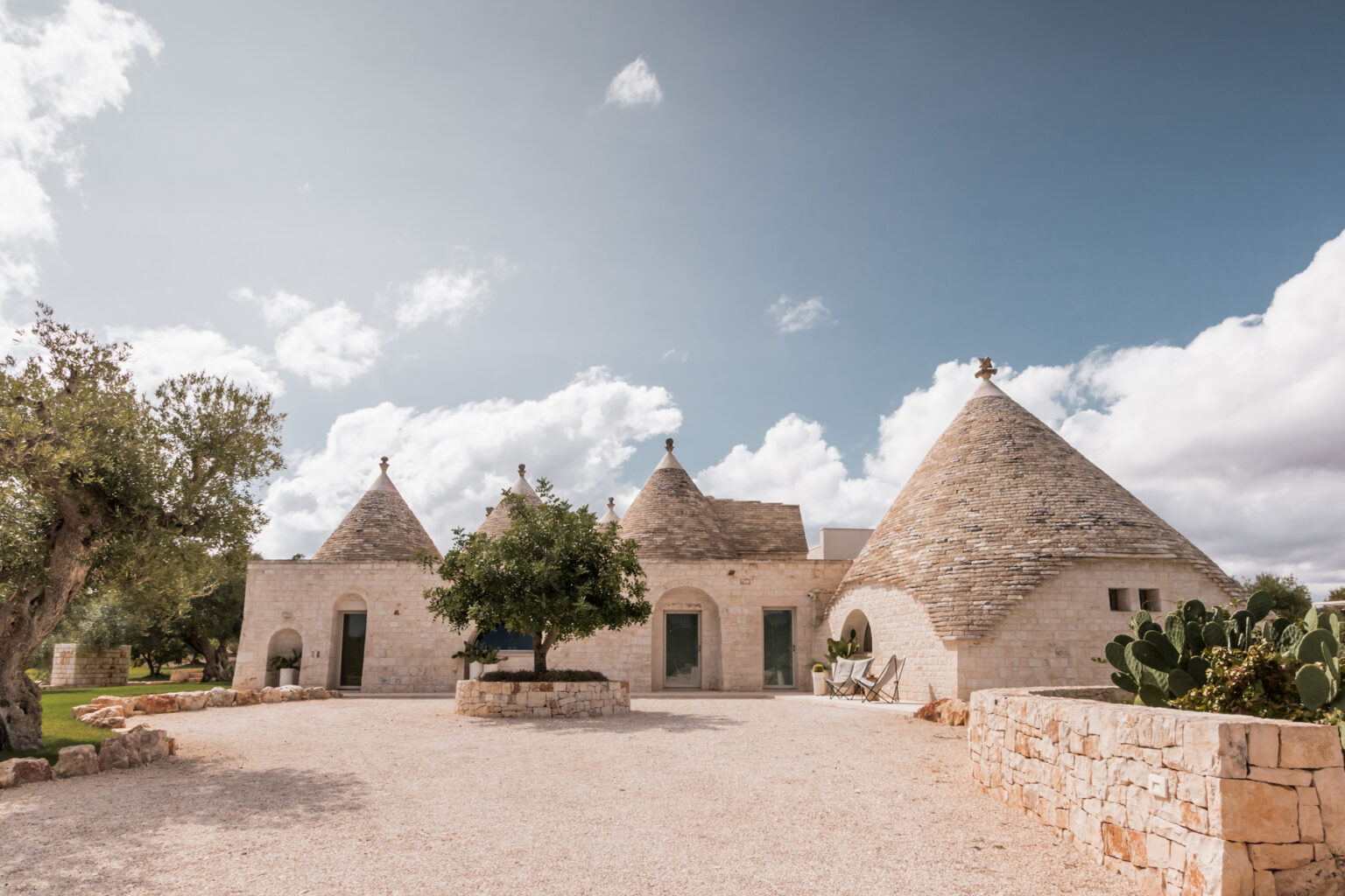Traditional trullo houses in Puglia, Italy, featuring conical stone roofs and surrounded by lush greenery under a partly cloudy sky. | Only on SopranoVillas