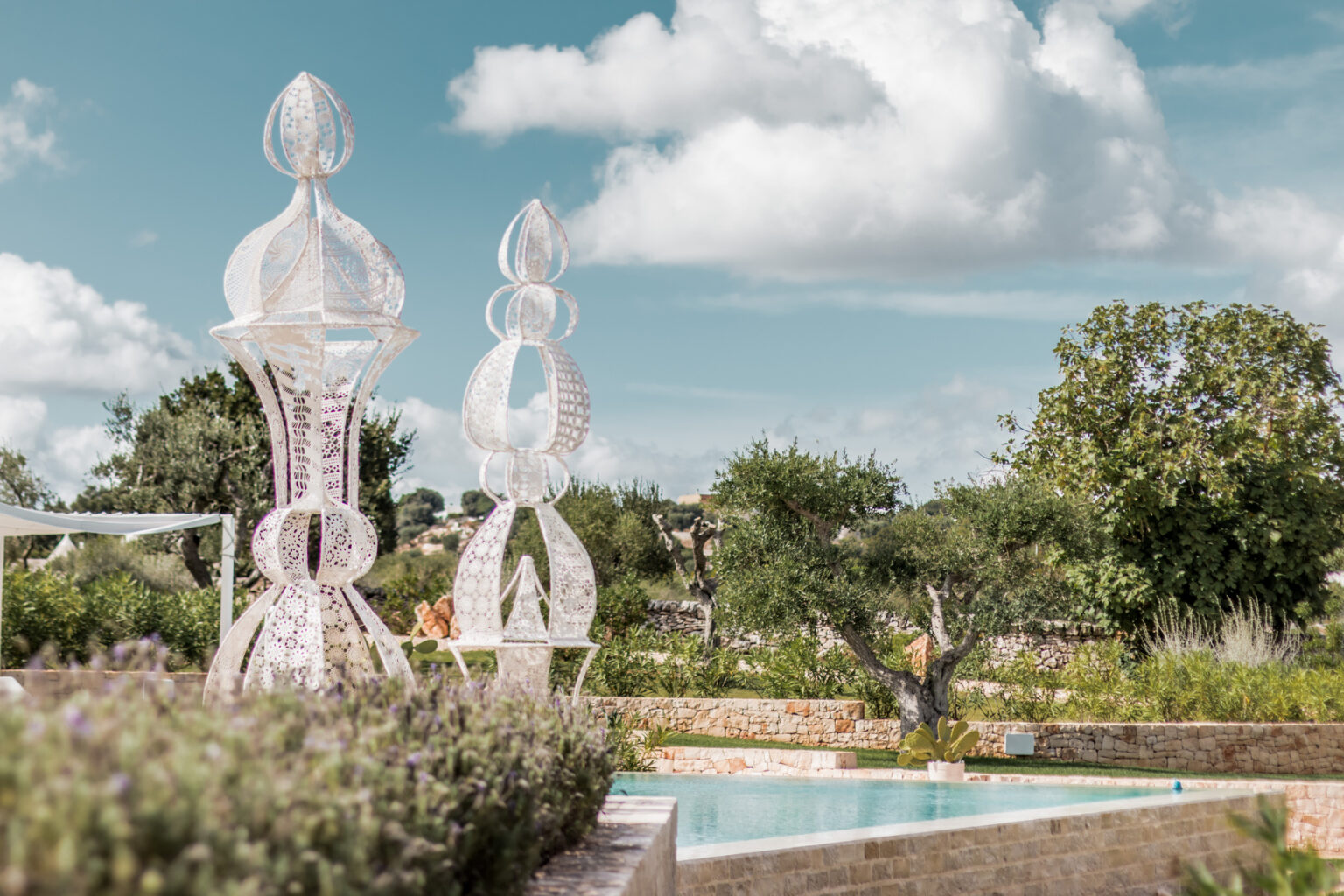 Elegant outdoor sculptures near a serene swimming pool, surrounded by lush greenery and a clear blue sky with fluffy clouds. Perfect for showcasing garden art and tranquil landscapes. | Only on SopranoVillas