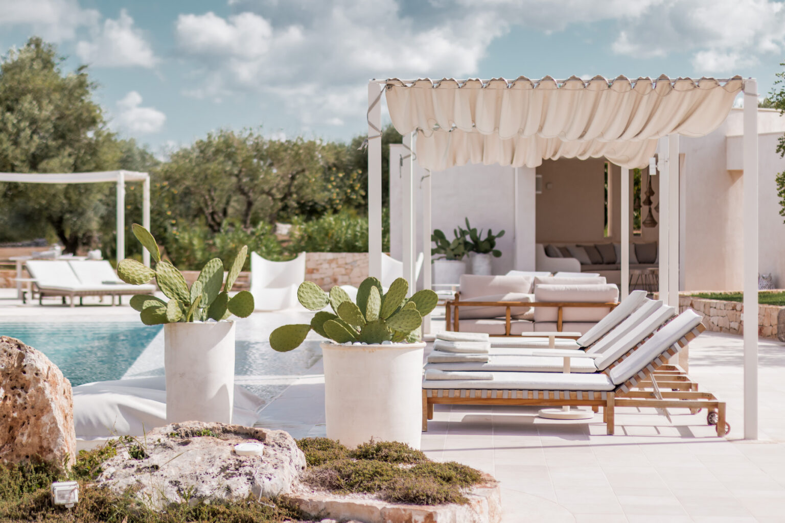 Luxurious outdoor pool area featuring modern lounge chairs, elegant cabana, and potted plants, surrounded by lush greenery and a clear blue sky, perfect for relaxation and leisure. | Only on SopranoVillas