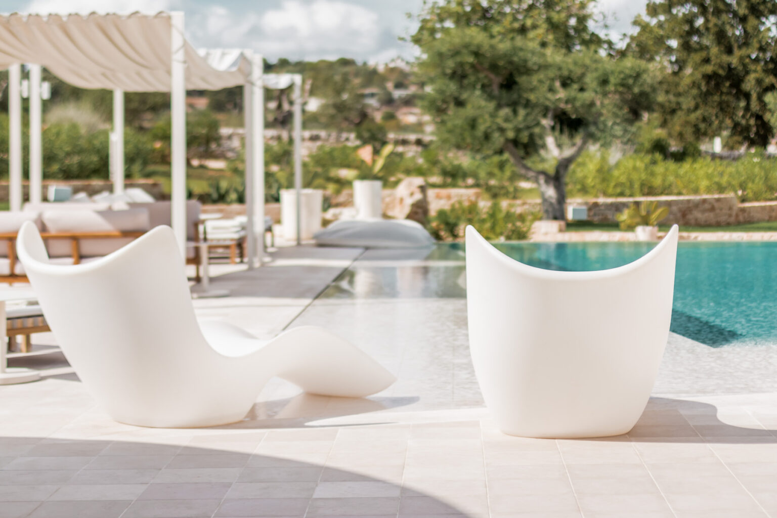 Modern outdoor lounge chairs by a swimming pool, featuring a sleek white design and surrounded by lush greenery. Perfect for relaxing in a luxurious setting. | Only on SopranoVillas