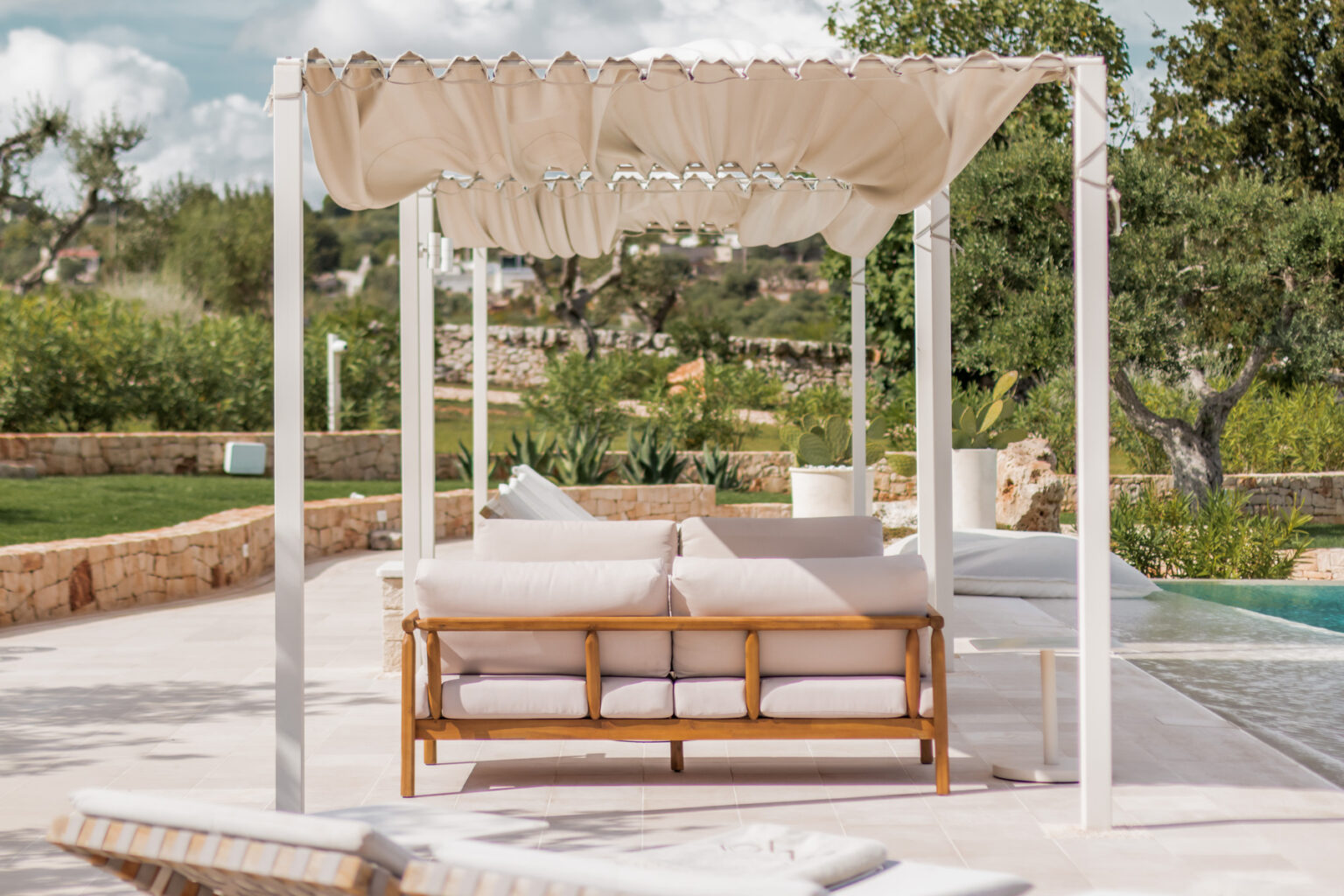 Cozy outdoor lounge area featuring a wooden sofa with soft cushions, shaded by a cream-colored canopy, surrounded by lush greenery and a swimming pool in a serene setting. | Only on SopranoVillas