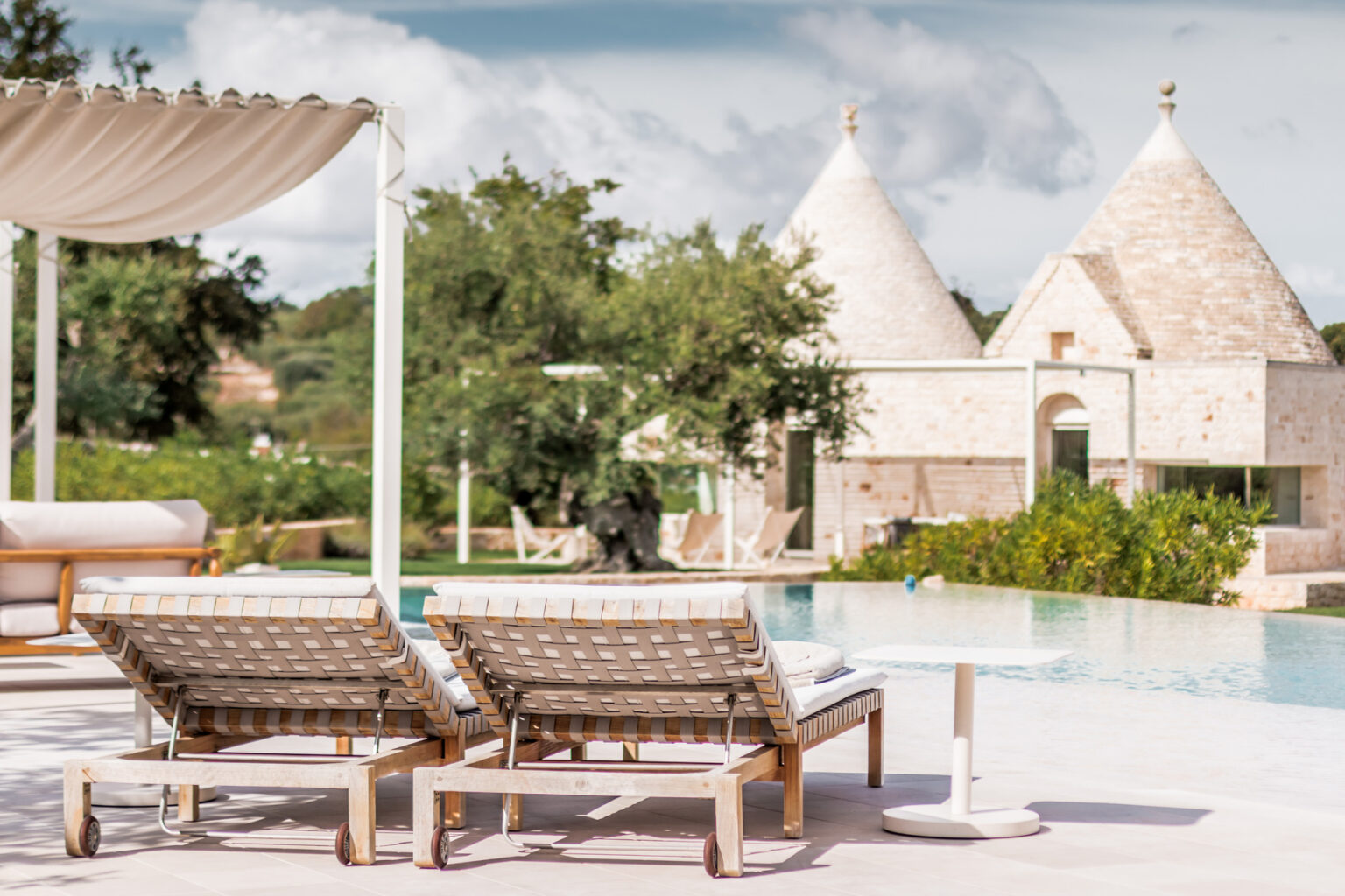 Lounge chairs beside a serene swimming pool with trullo-style buildings in the background, surrounded by lush greenery under a partly cloudy sky. Ideal for luxury vacation or relaxing getaway settings. | Only on SopranoVillas