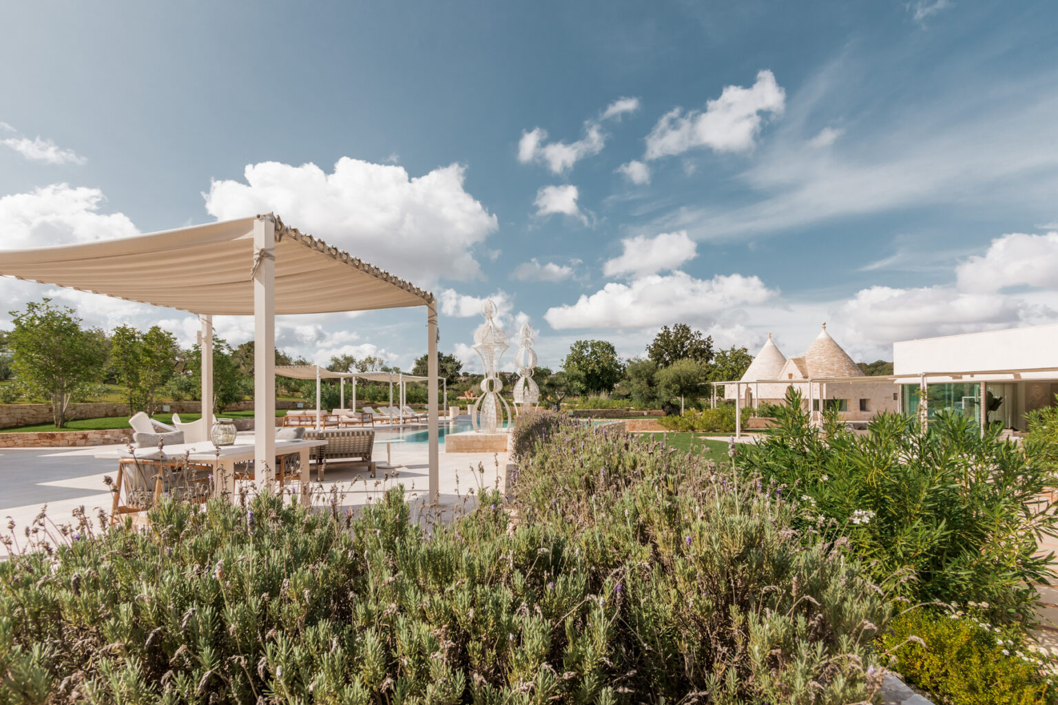 Luxurious outdoor patio area featuring a shaded seating arrangement, a swimming pool, and traditional trulli houses, surrounded by lush greenery and blooming lavender under a bright blue sky with fluffy white clouds. | Only on SopranoVillas