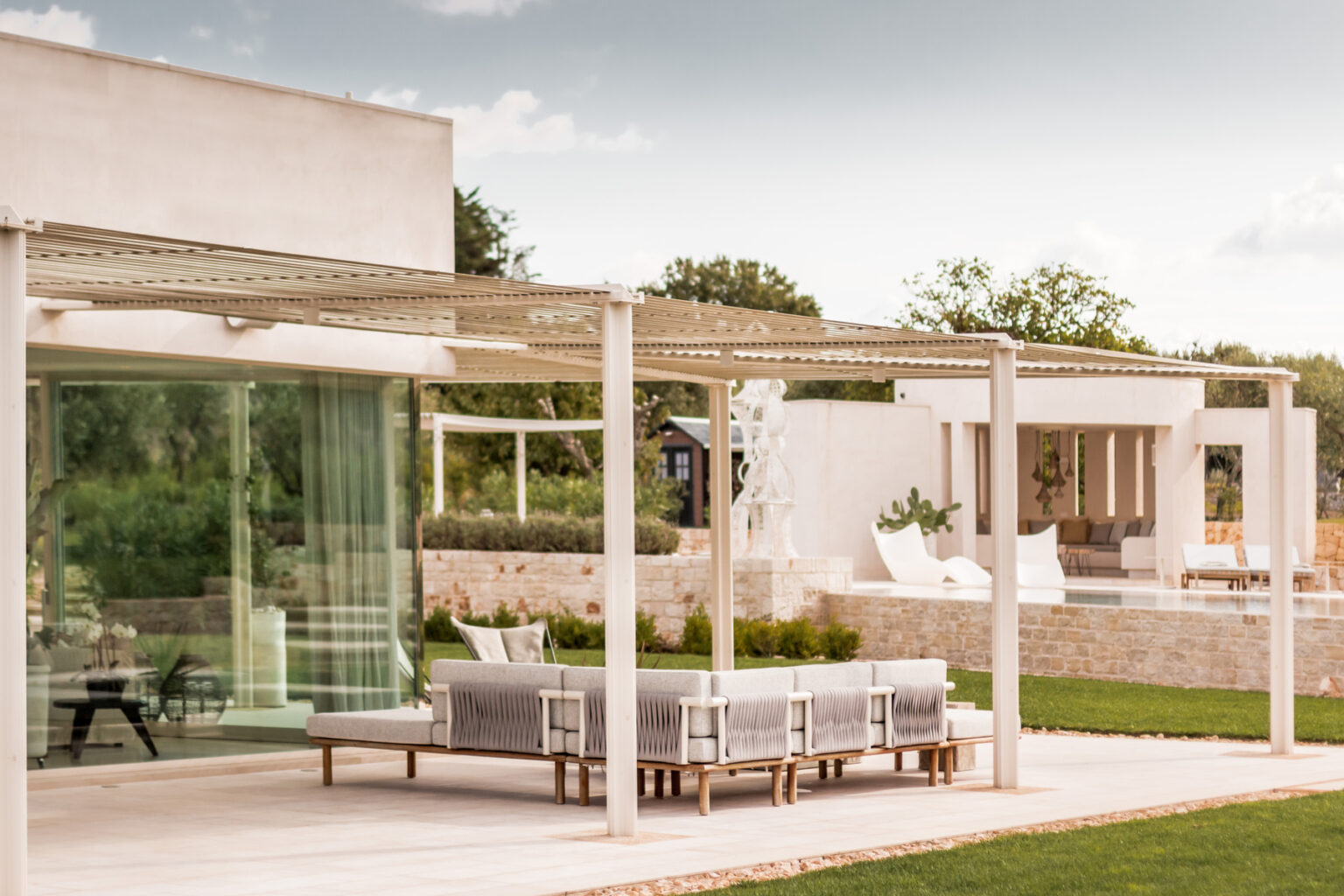 Modern outdoor patio with a gray seating area and pergola, featuring a spacious garden and contemporary architecture in the background. Ideal for relaxation and entertaining. | Only on SopranoVillas