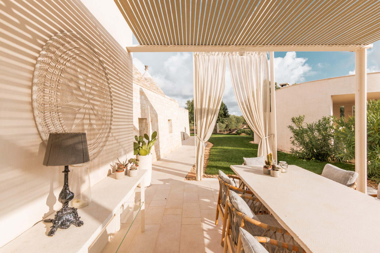 Elegant outdoor dining area featuring a long table with stylish chairs, shaded by a wooden pergola and flowing curtains, surrounded by lush greenery and a bright sky. Perfect for al fresco dining and entertaining. | Only on SopranoVillas