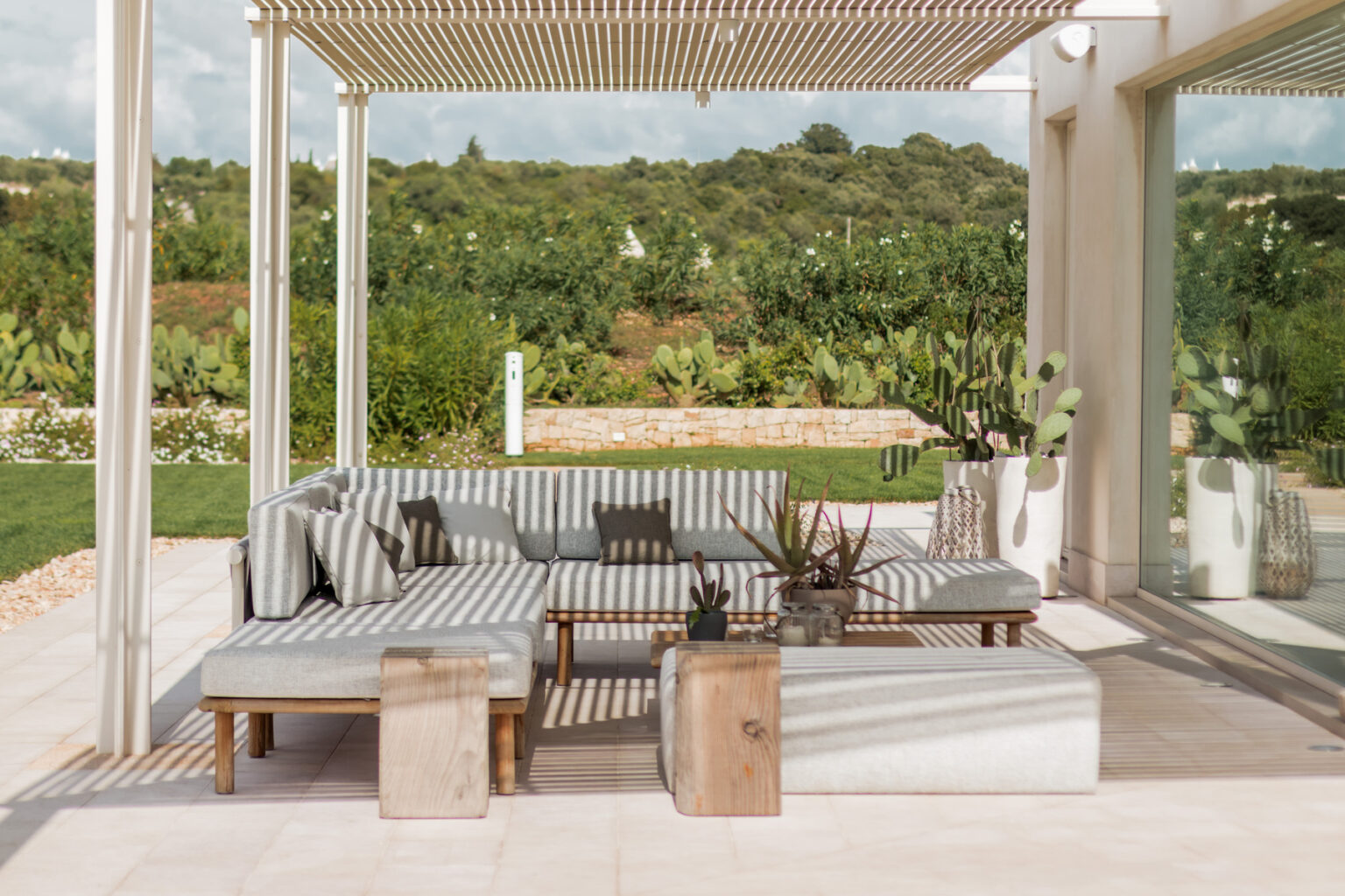 Modern outdoor living space featuring a striped sectional sofa, natural wood coffee tables, and potted plants, set against a lush green landscape under a pergola. Ideal for relaxation and entertaining. | Only on SopranoVillas