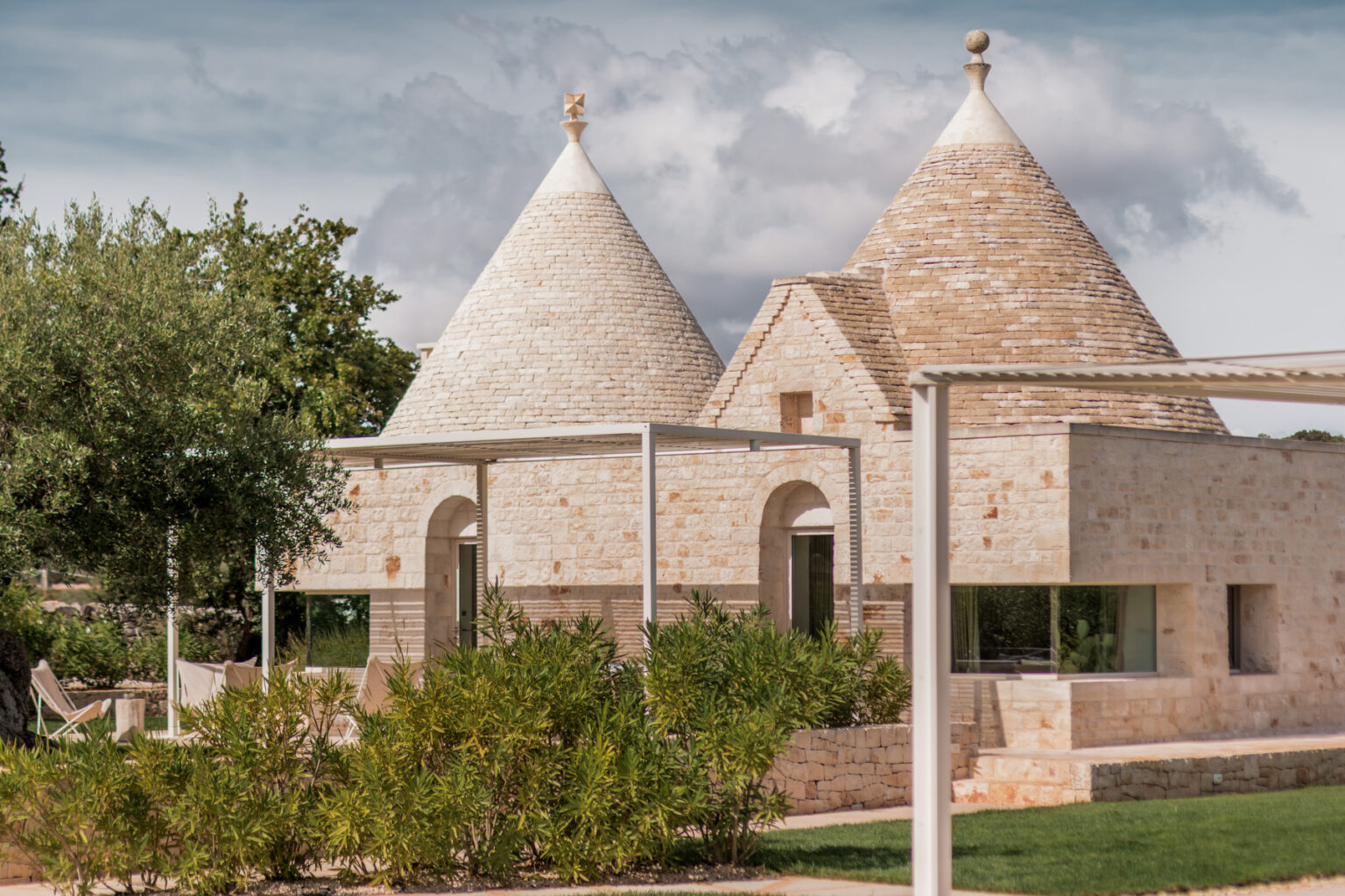 Traditional trulli houses in Apulia, Italy, showcasing conical roofs and natural stone construction, surrounded by greenery and a modern outdoor space. | Only on SopranoVillas