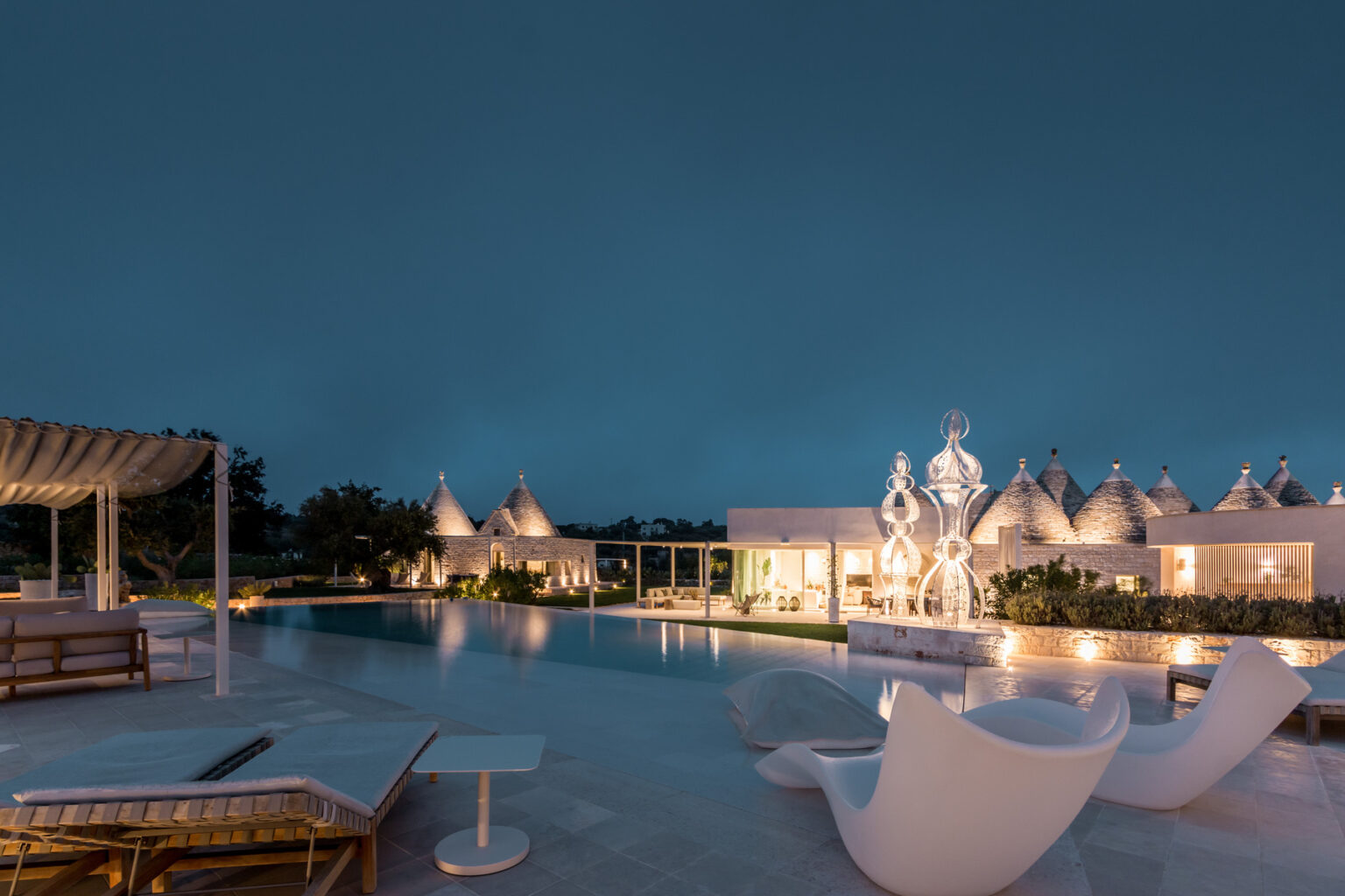 Modern resort at night featuring a tranquil pool, stylish lounge chairs, and unique architectural structures in the background, surrounded by lush greenery. | Only on SopranoVillas