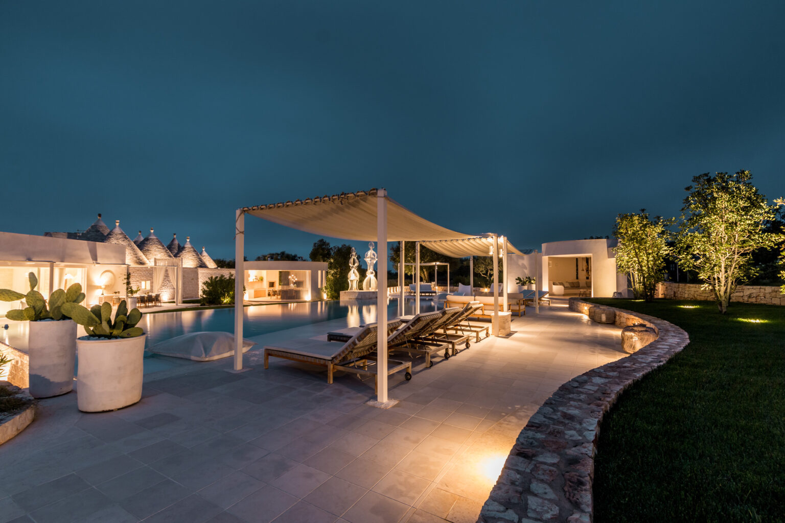 Modern outdoor pool area at night featuring lounge chairs, elegant lighting, and landscaped greenery, creating a serene and inviting atmosphere. | Only on SopranoVillas