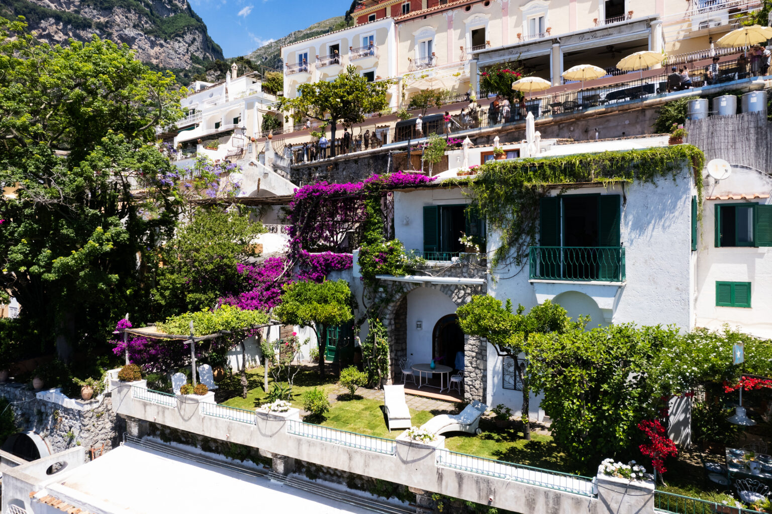 Charming hillside villa in Positano, Italy, surrounded by vibrant bougainvillea and lush greenery. The image showcases a sunny outdoor garden with loungers and a stone patio, nestled among traditional Mediterranean architecture and terraces. Ideal for travelers seeking a picturesque coastal getaway. | Only on SopranoVillas