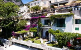 Charming hillside villa in Positano, Italy, surrounded by vibrant bougainvillea and lush greenery. The image showcases a sunny outdoor garden with loungers and a stone patio, nestled among traditional Mediterranean architecture and terraces. Ideal for travelers seeking a picturesque coastal getaway.