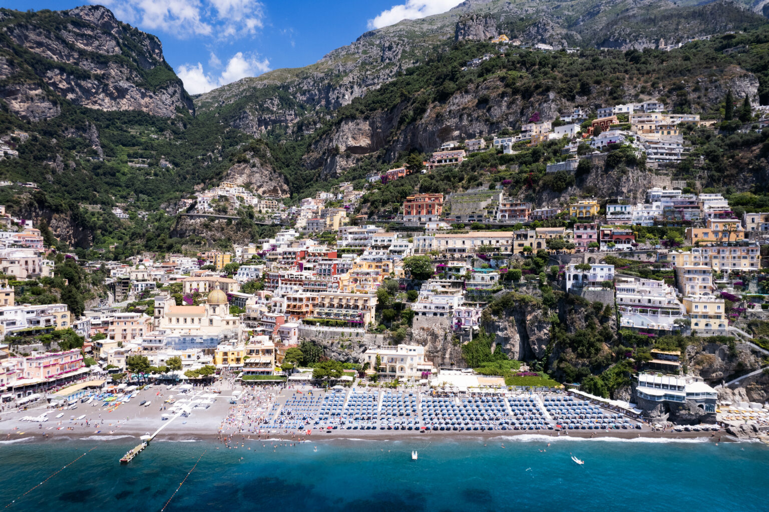 Aerial view of Positano, Italy, showcasing colorful cliffside buildings, sandy beach with sun loungers, and the stunning Amalfi Coast landscape in the background. | Only on SopranoVillas