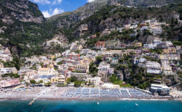 Aerial view of Positano, Italy, showcasing colorful cliffside buildings, sandy beach with sun loungers, and the stunning Amalfi Coast landscape in the background.